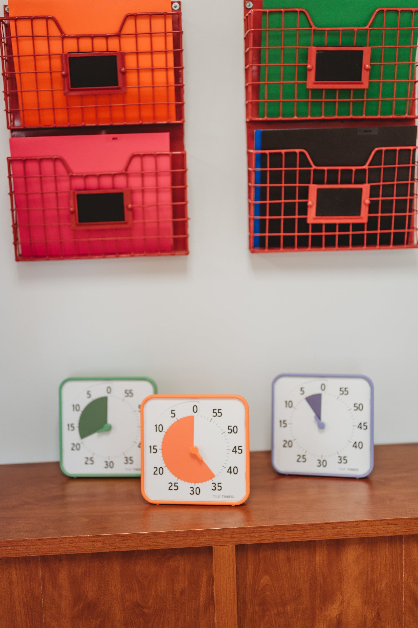The Time Timer Original (8 inch) Learning Center Classroom Set by Time Timer features three timers with green, orange, and purple borders, perfect for classroom time management. Four red and orange wall storage baskets are displayed above a wooden cabinet.