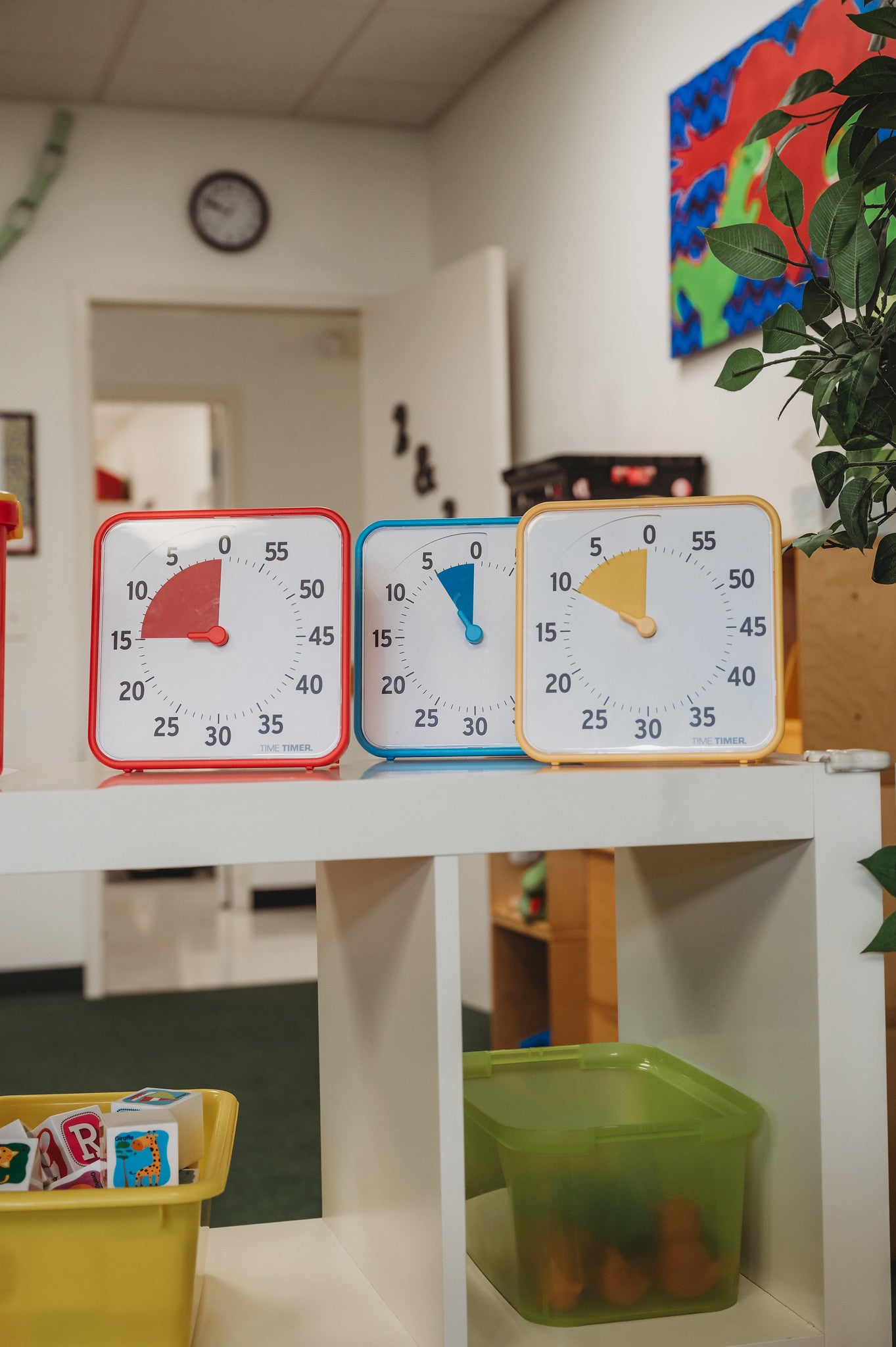 The Time Timer Original (8 inch) Learning Center Classroom Set by Time Timer in primary colors sits on a classroom shelf among toys and bins, with vibrant decorations and a wall clock, creating an engaging space for time management.
