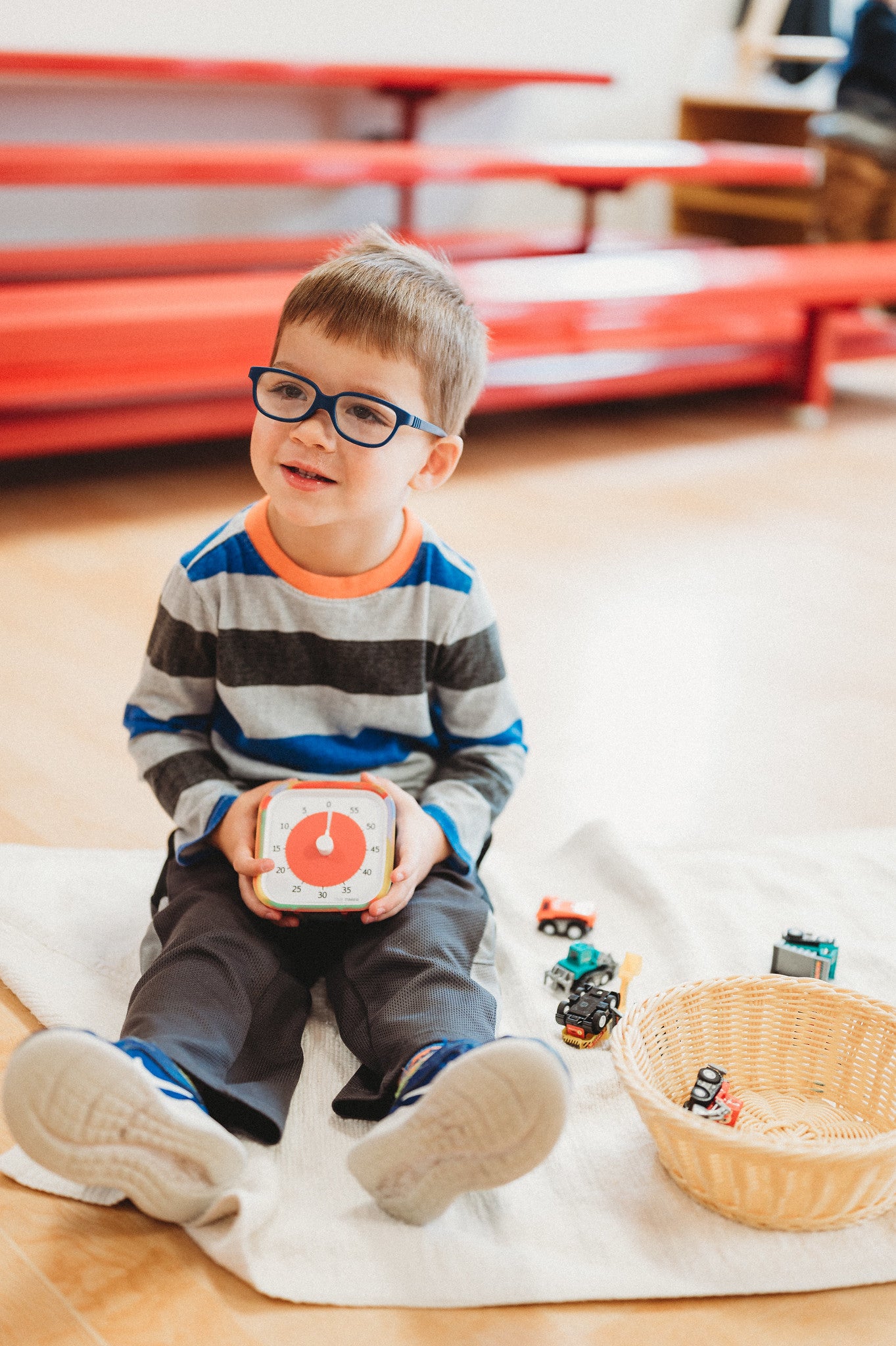 A young boy with glasses, in a striped shirt and gray pants, sits on the floor holding the Time Timer MOD [Tie Dye] (60 min) by Time Timer. A basket and toy cars are nearby on a white mat, with red benches in the background.