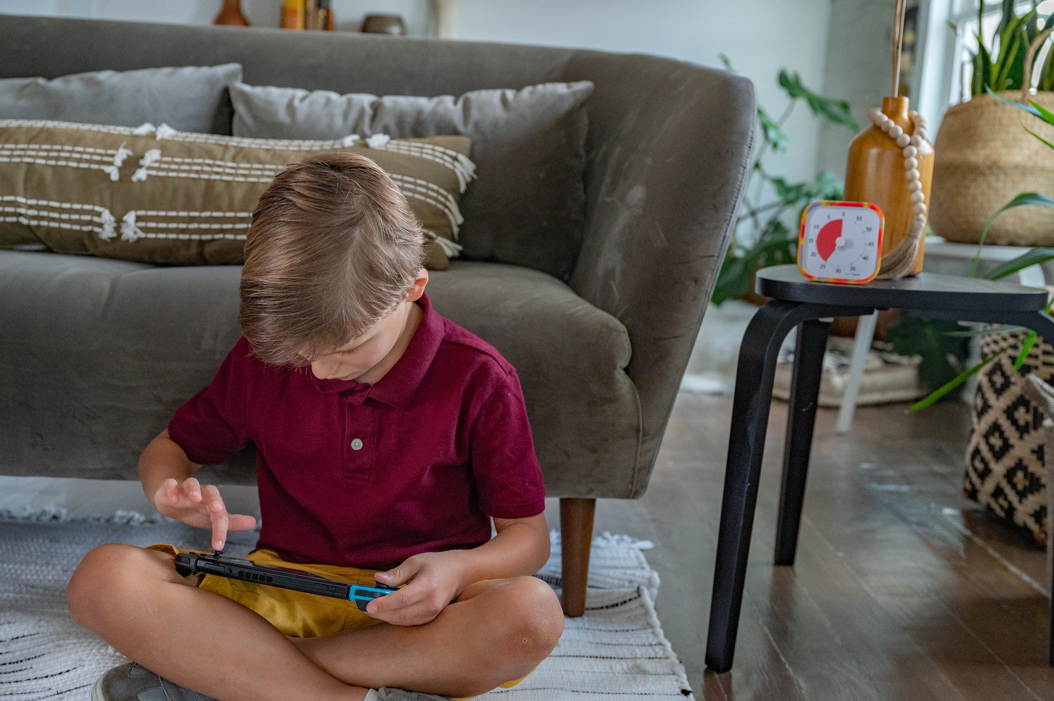 A young boy sits cross-legged on a rug in front of a couch, focused on a handheld game with a silicone case. Nearby, the Time Timer MOD [Tie Dye] (60 min) by Time Timer and a vase sit on a small table, with plants and home decor in the background.
