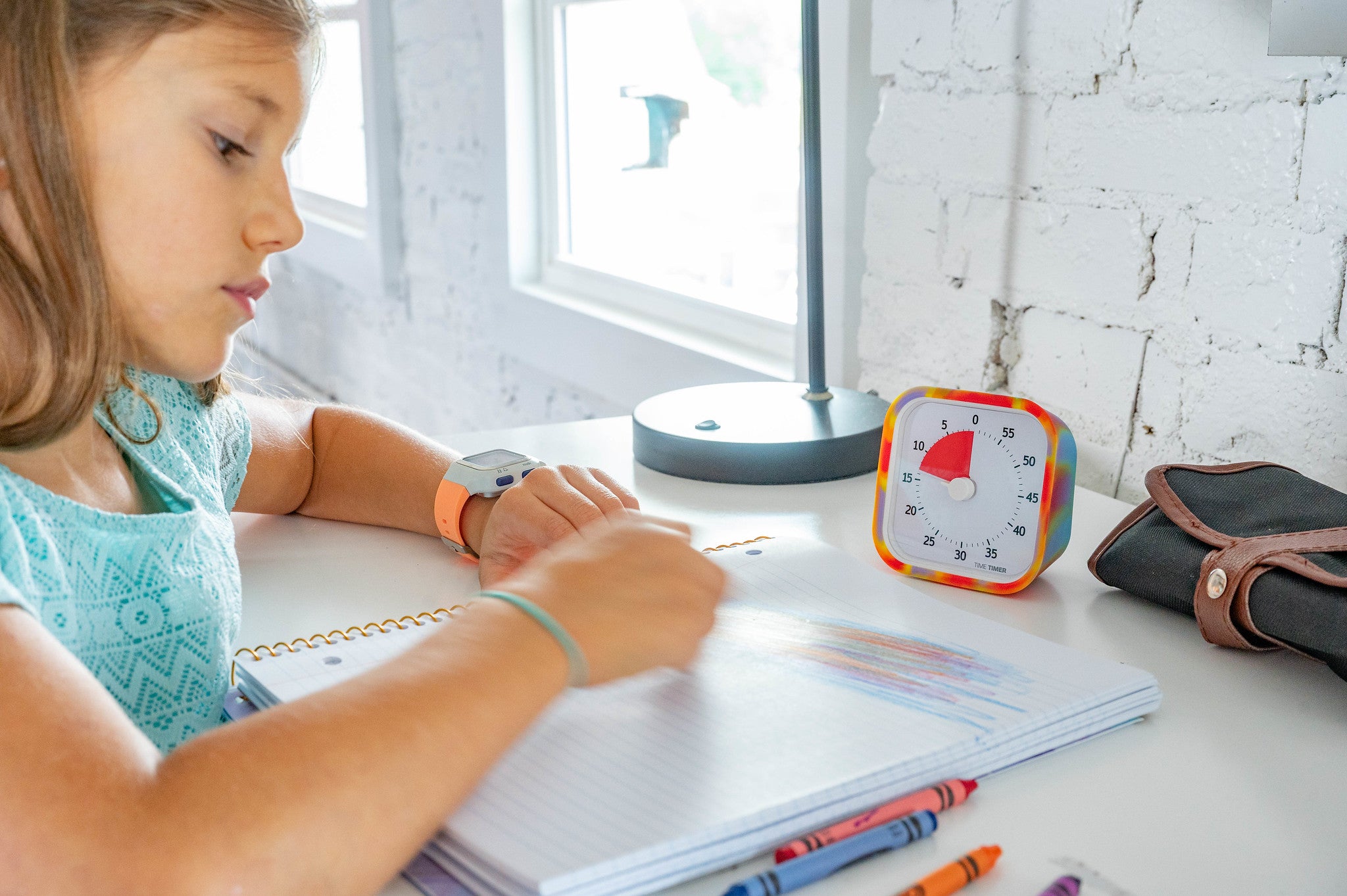A young girl draws with colored crayons in a notebook at a white desk. On the desk is the Time Timer MOD [Tie Dye] (60 min) from Time Timer, along with a lamp, pencil case, more crayons, and a white brick wall background.