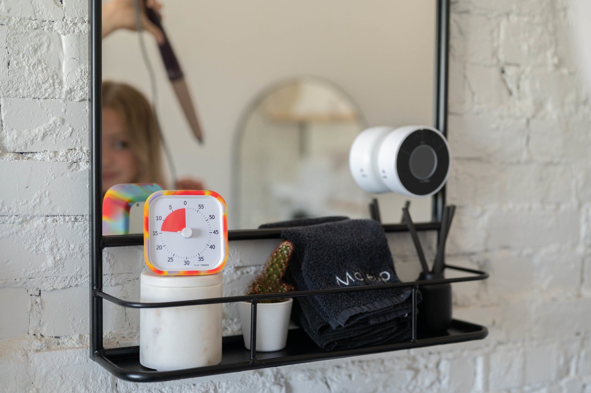 A Time Timer MOD [Tie Dye] by Time Timer in a silicone case, a white jar, dark towels, and toiletries sit on a bathroom shelf near a mirror reflecting a child brushing their hair.