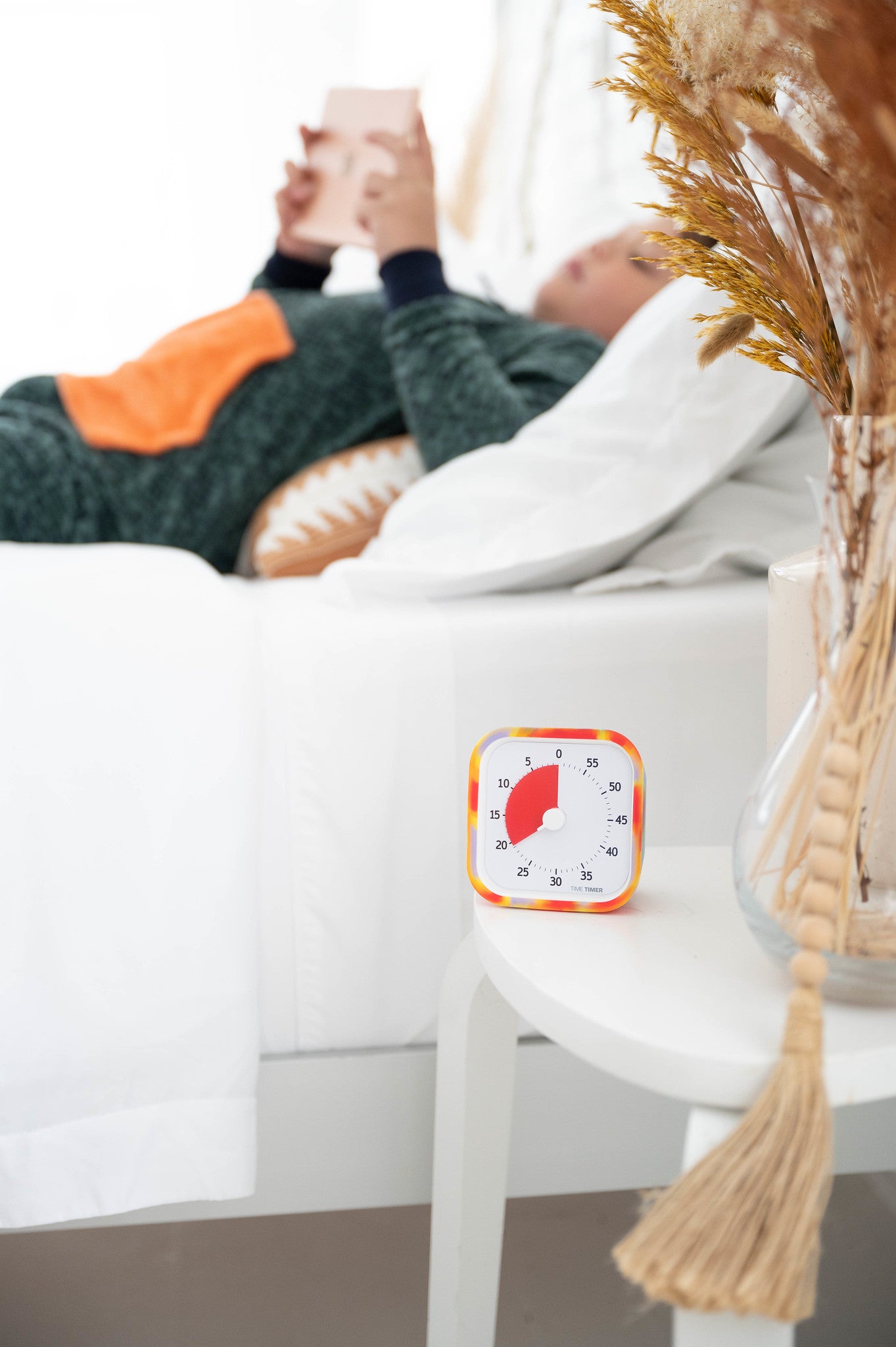 A person lies on a bed reading, partially covered by a blanket. On the white bedside table sits a Time Timer MOD [Tie Dye] (60 min) by Time Timer in its silicone case, next to a vase with dried plants.
