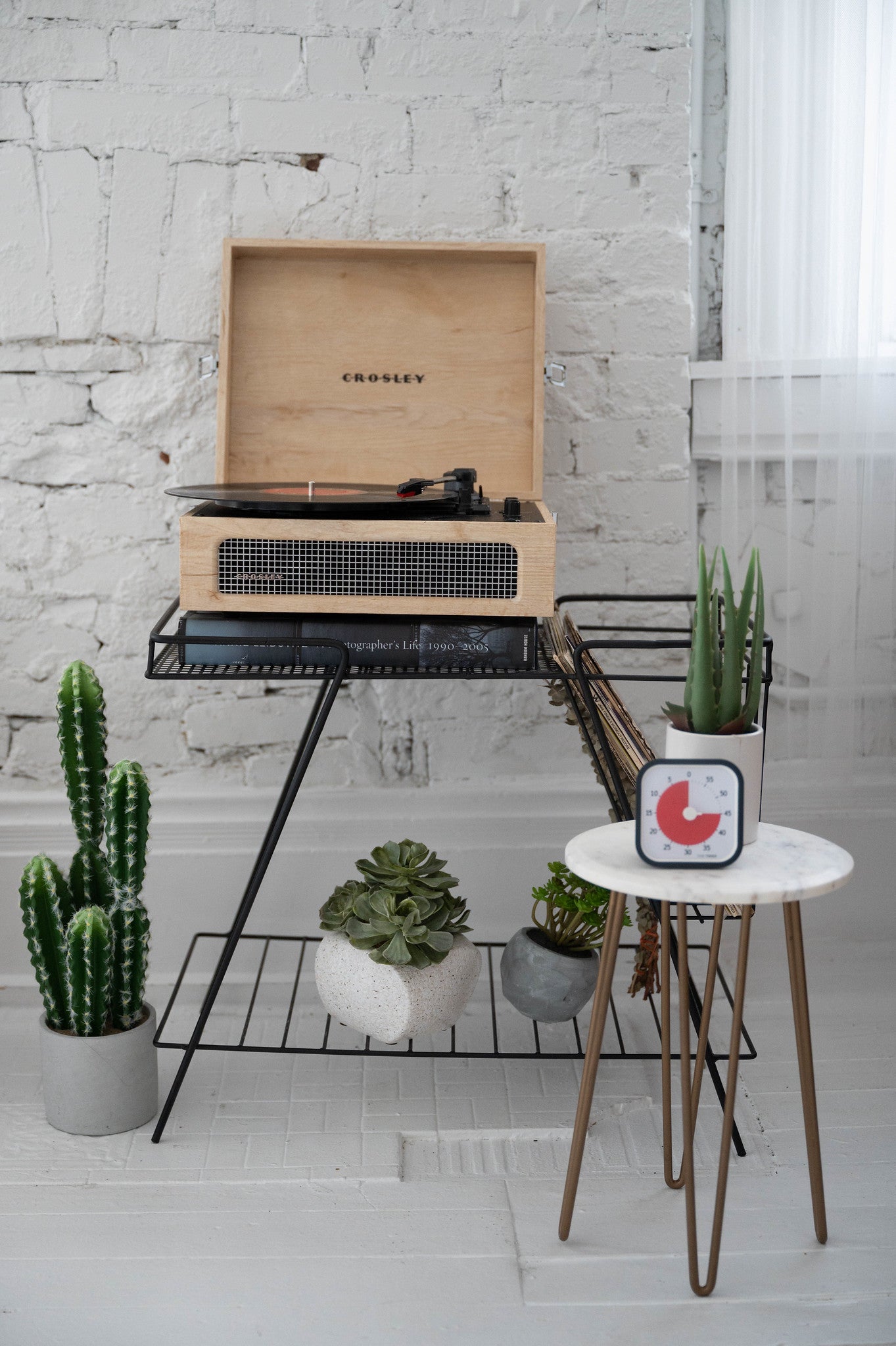 A Crosley record player rests on a black metal stand with vinyl records, surrounded by potted cacti. On a small round table is the Time Timer MOD [Charcoal] (60 min) by Time Timer and a plant, set against a white brick wall and sheer curtains.