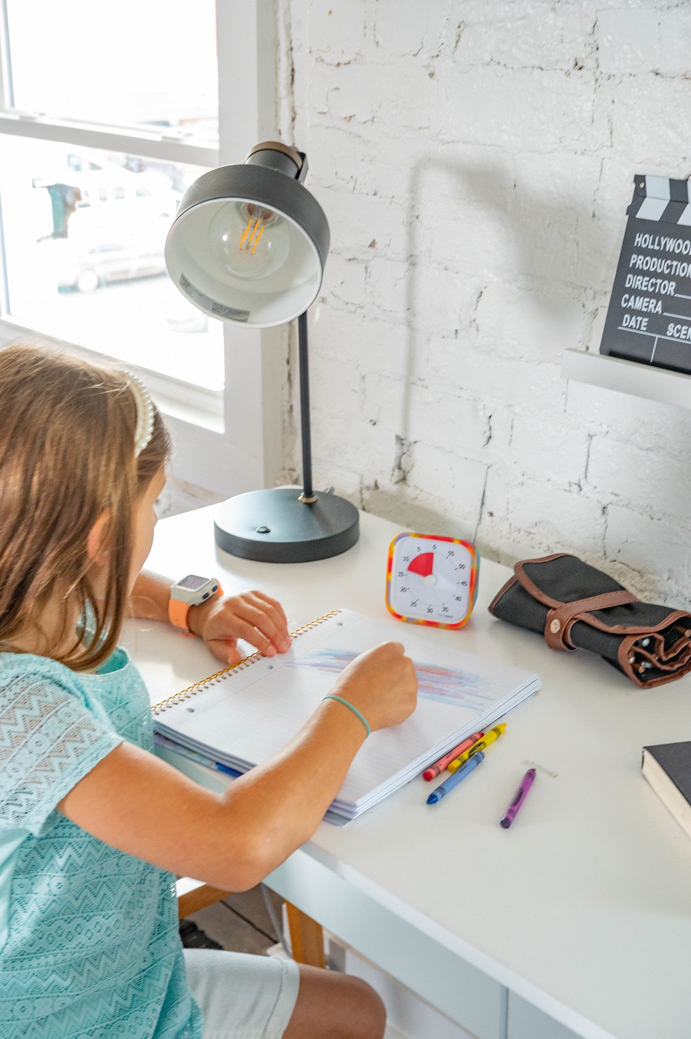 A young girl draws with colored pencils at a white desk, using a lamp, a clock, and the Time Timer MOD [Tie Dye] by Time Timer for effective time management as she focuses on her artwork in the bright room.