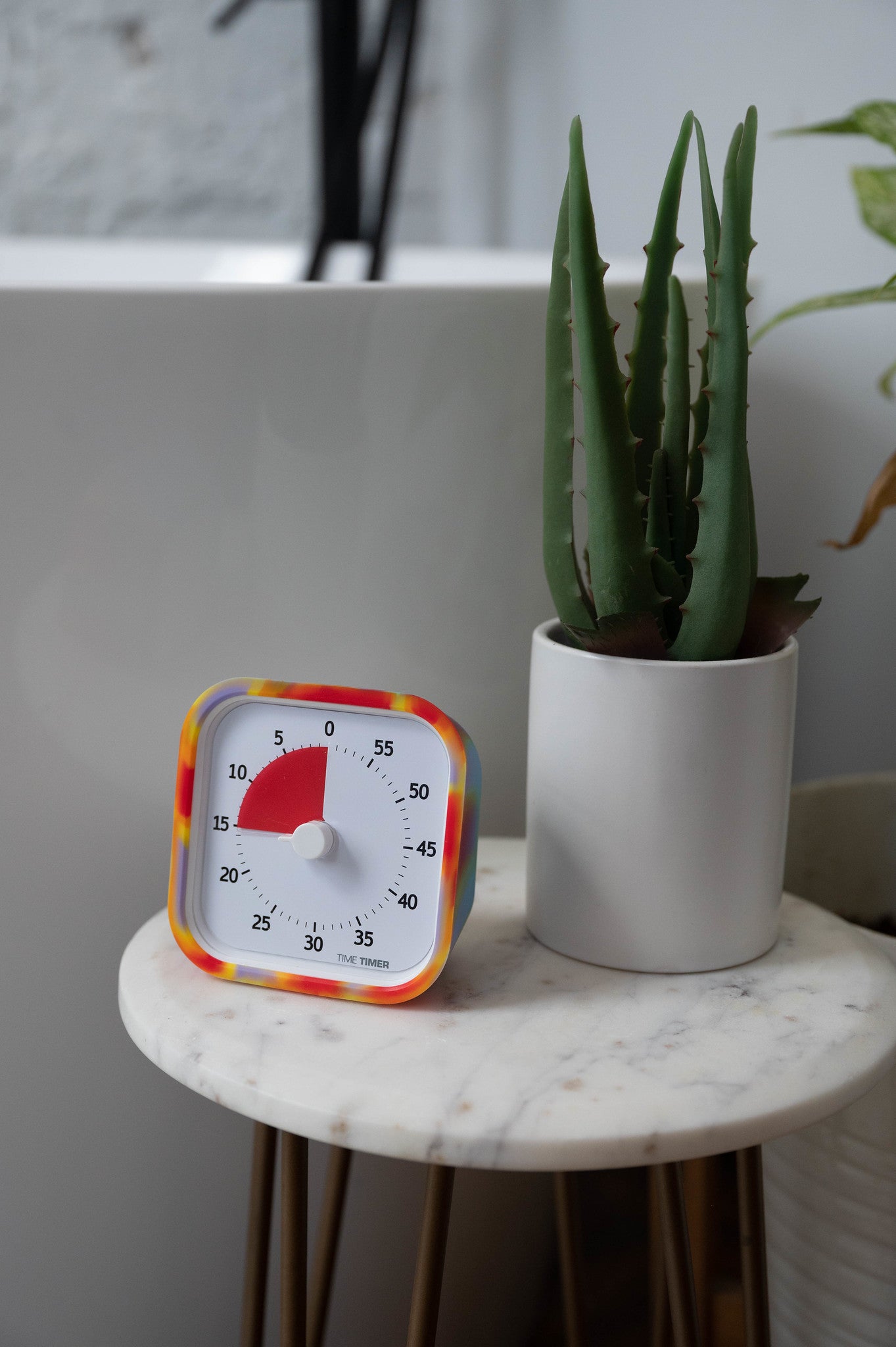 The Time Timer MOD [Tie Dye] (60 min) by Time Timer, featuring a red countdown display and silicone case, rests on a round marble-topped table beside a cactus, doubling as a practical time management tool and stylish decor.