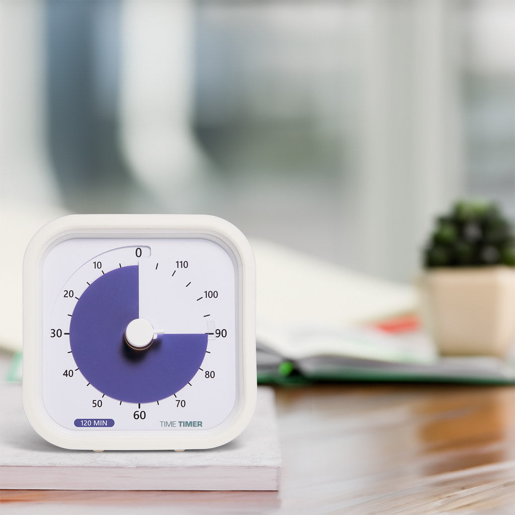 A white Time Timer MOD Education (120 min) by Time Timer sits on a desk for visual time management, with open books and a small potted plant blurred in the background.
