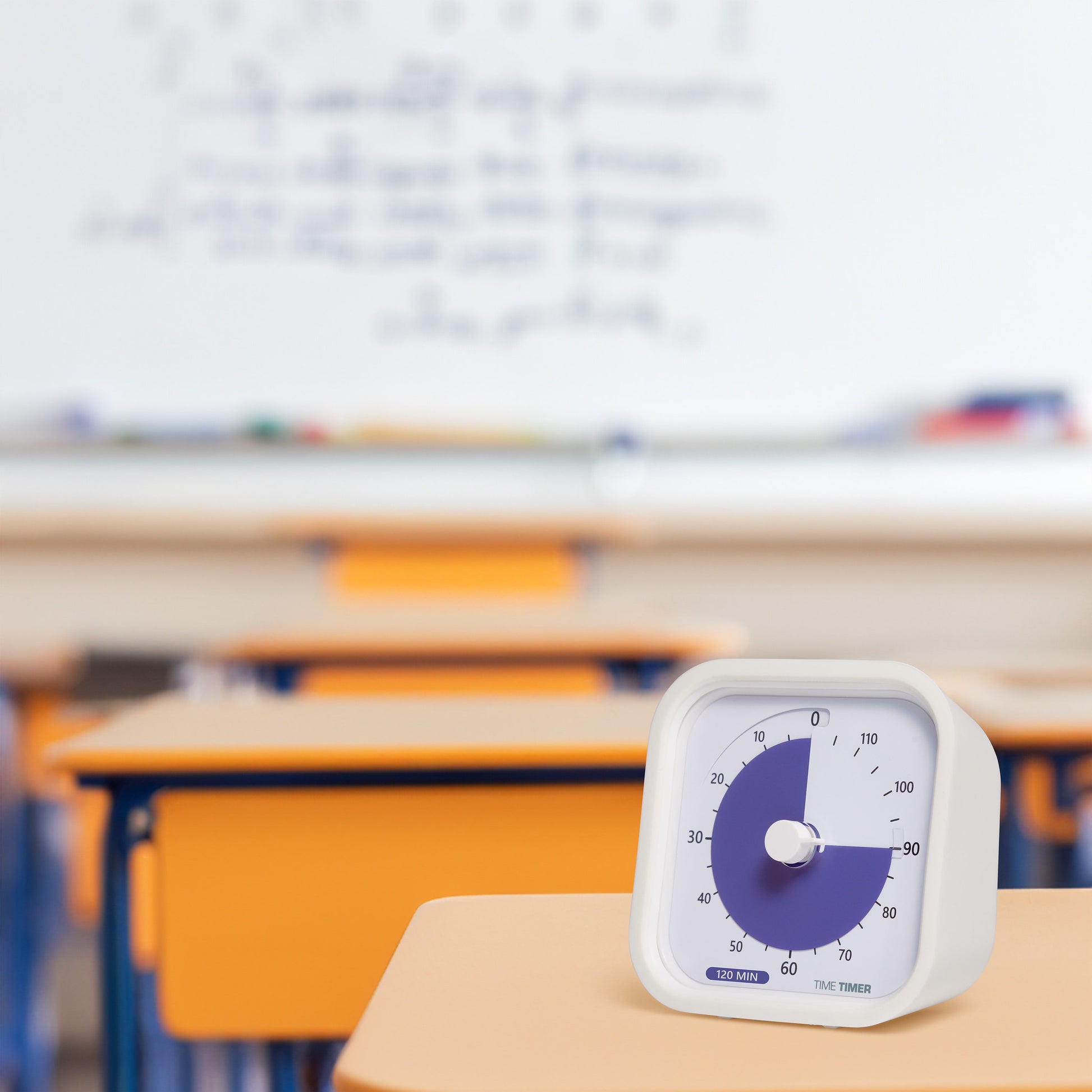 A Time Timer MOD Education (120 min) by Time Timer, featuring a purple disk, is placed on a school desk for visual time management. Blurred classroom desks and a whiteboard can be seen in the background.