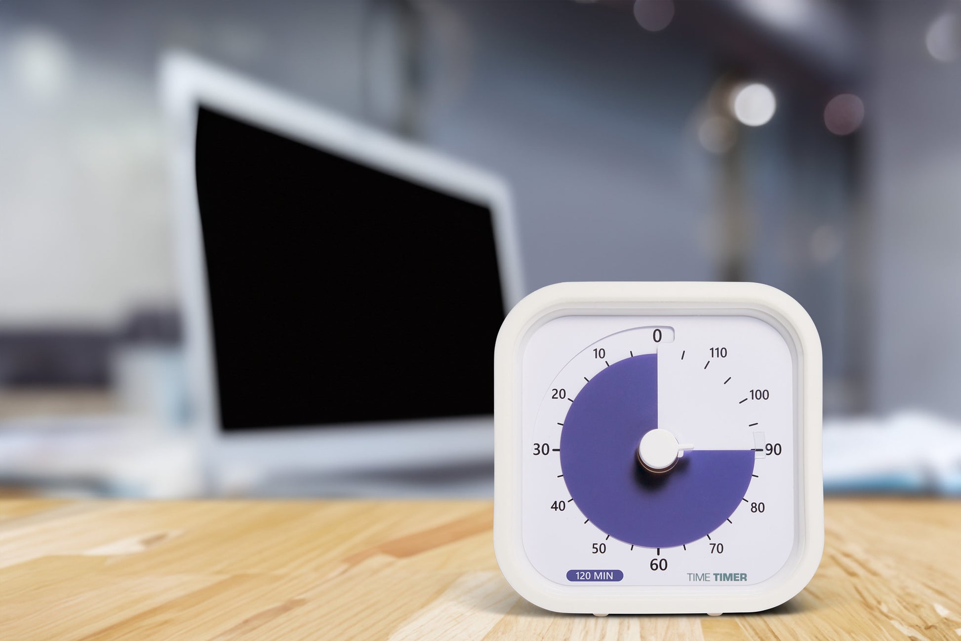 A Time Timer MOD Education (120 min) by Time Timer sits on a wooden desk, supporting visual time management. A blurred laptop and office background complete the scene.