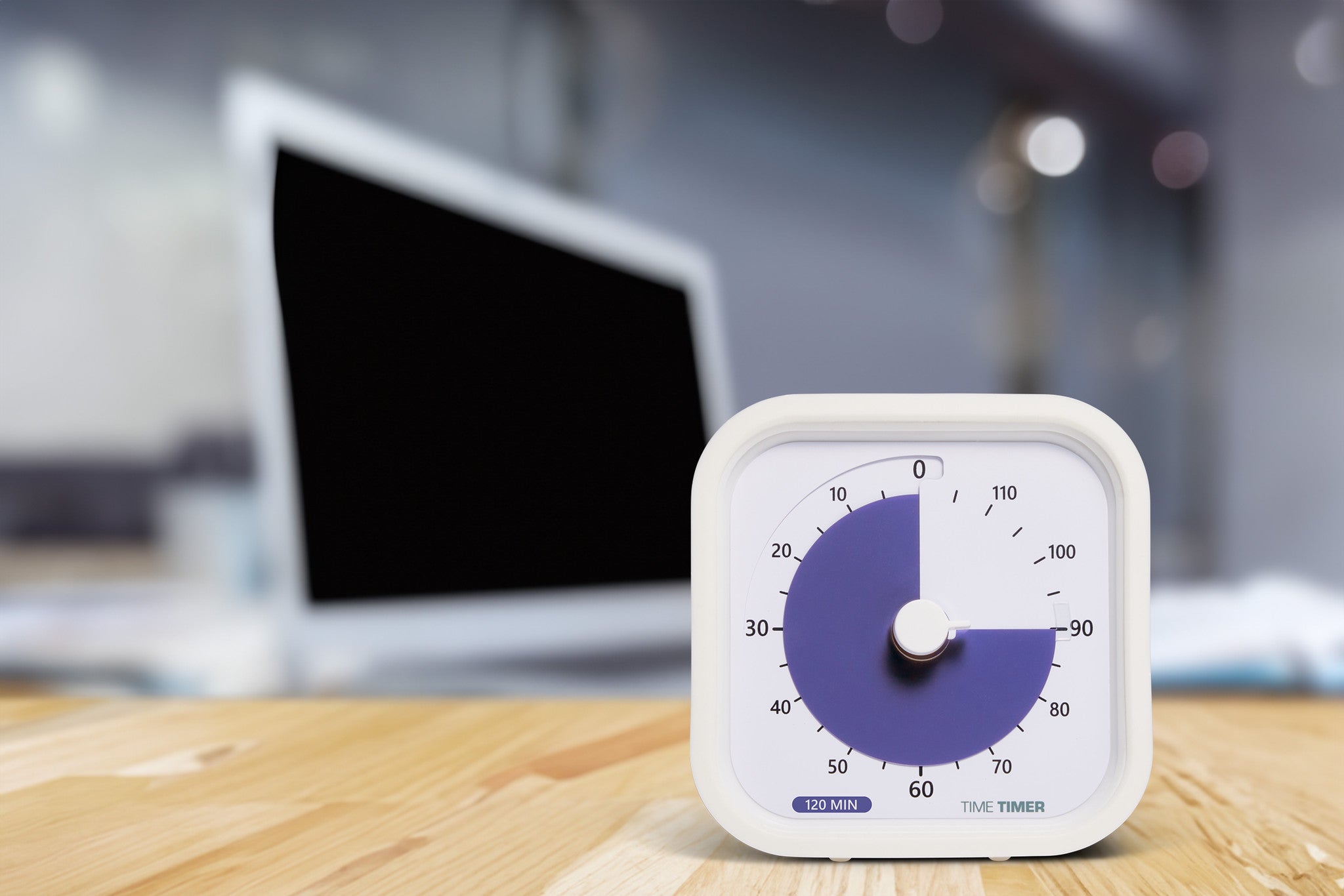A Time Timer MOD Education (120 min) by Time Timer sits on a wooden desk, supporting visual time management. A blurred laptop and office background complete the scene.