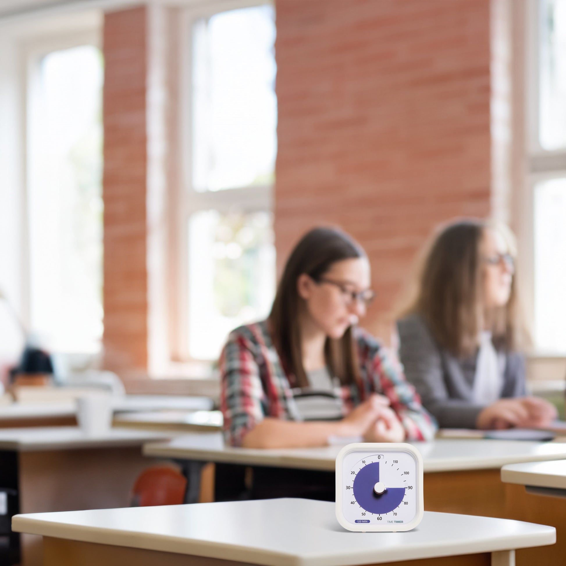 In a sunlit classroom, two students sit at desks in the background. A Time Timer MOD Education (120 min) by Time Timer with a blue dial is in focus on a desk, supporting visual time management.
