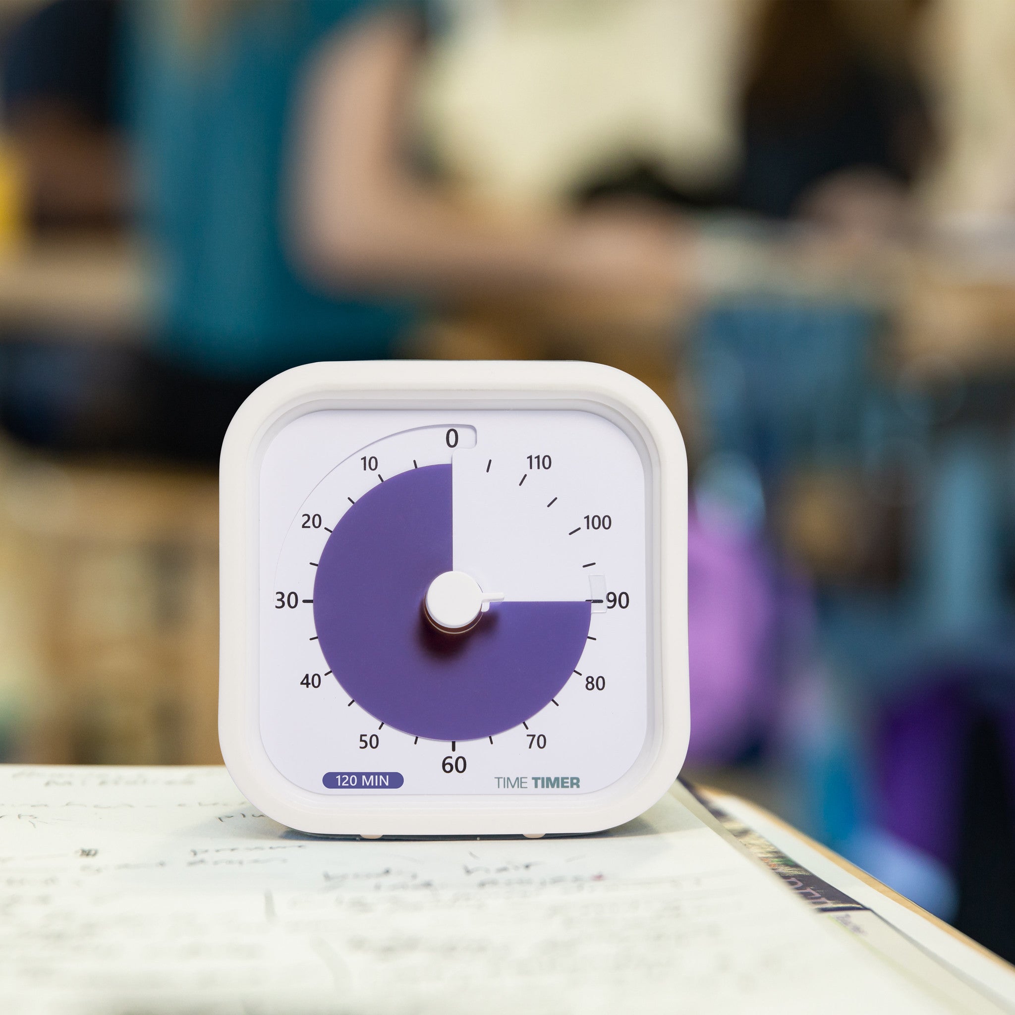 A Time Timer MOD Education (120 min) by Time Timer, in white, displays 30 minutes left atop a notebook, with a blurred classroom and students in the background—ideal for visual time management.