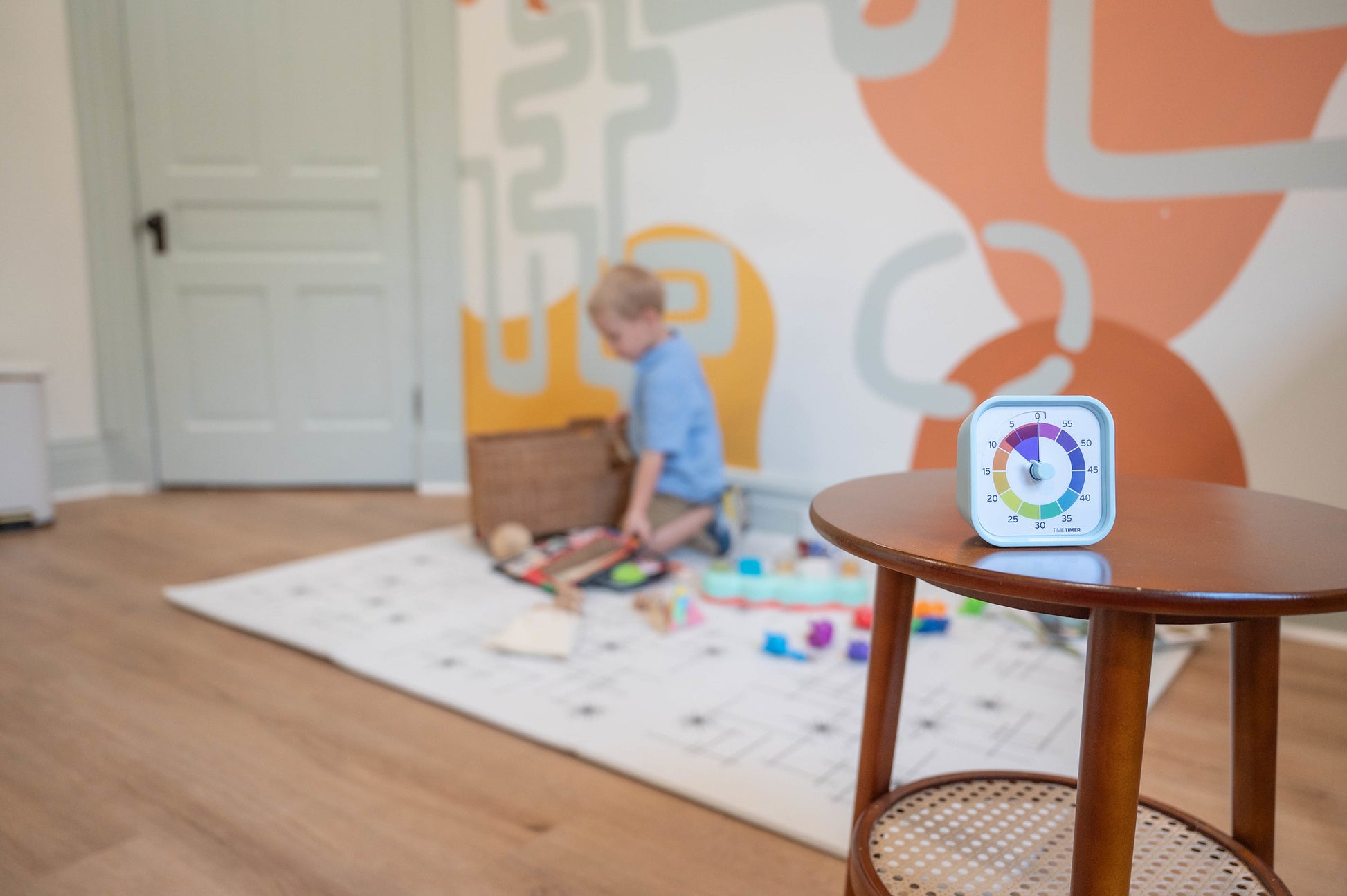 A Time Timer MOD - Home Edition [Rainbow Wheel] by Time Timer sits on a round wooden table, while a child plays with toys in a bright, colorful room—an inviting setting ideal for autism support.