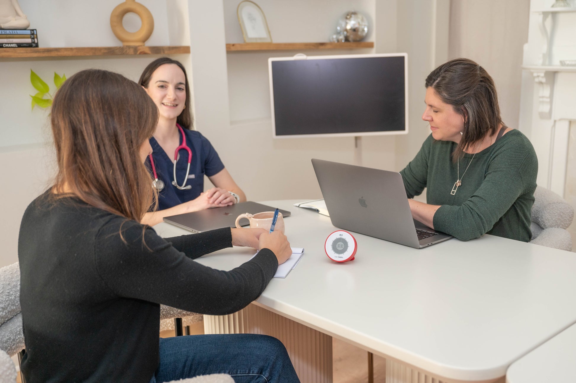 Three women sit around a table in an office. One wears scrubs with a stethoscope, another types on a laptop, and the third writes in a notebook. A computer monitor, small clock, and Time Timer Twist with Kickstand (Red) by Time Timer are on the table.