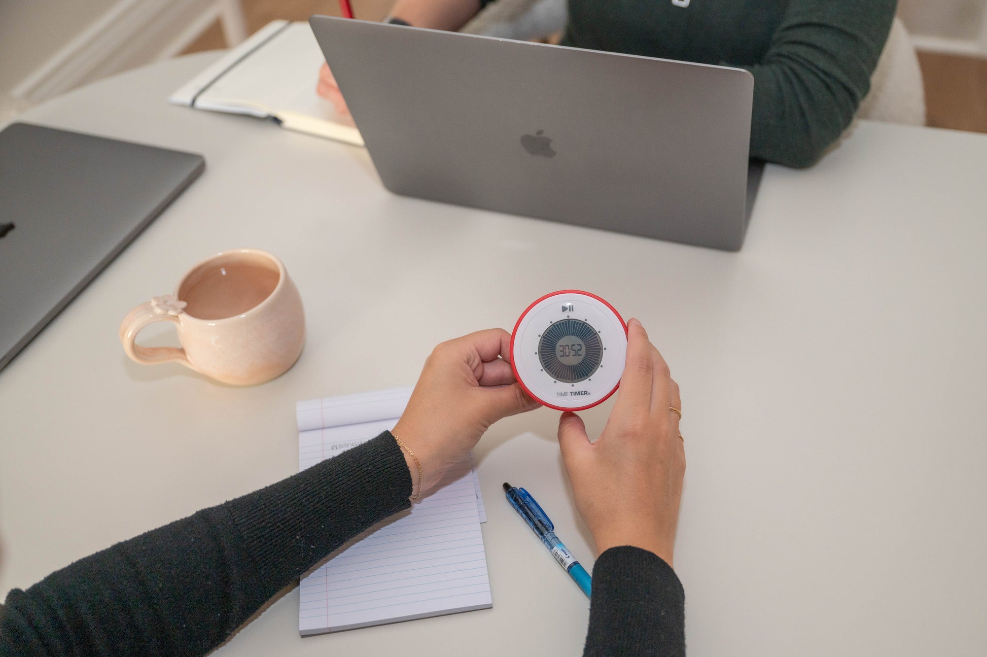 A person uses a red Time Timer Twist with Kickstand (Time Timer brand), set to 25 minutes, on a desk with a notepad, pen, mug, and open laptop. Another person sits across from them working on their own laptop.