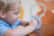 A young child in a blue shirt sits at a table, turning the dial on the Time Timer MOD - Home Edition [Rainbow Wheel] (60 min) by Time Timer. Abstract soft orange and beige shapes in the background create a calming, autism-friendly scene.