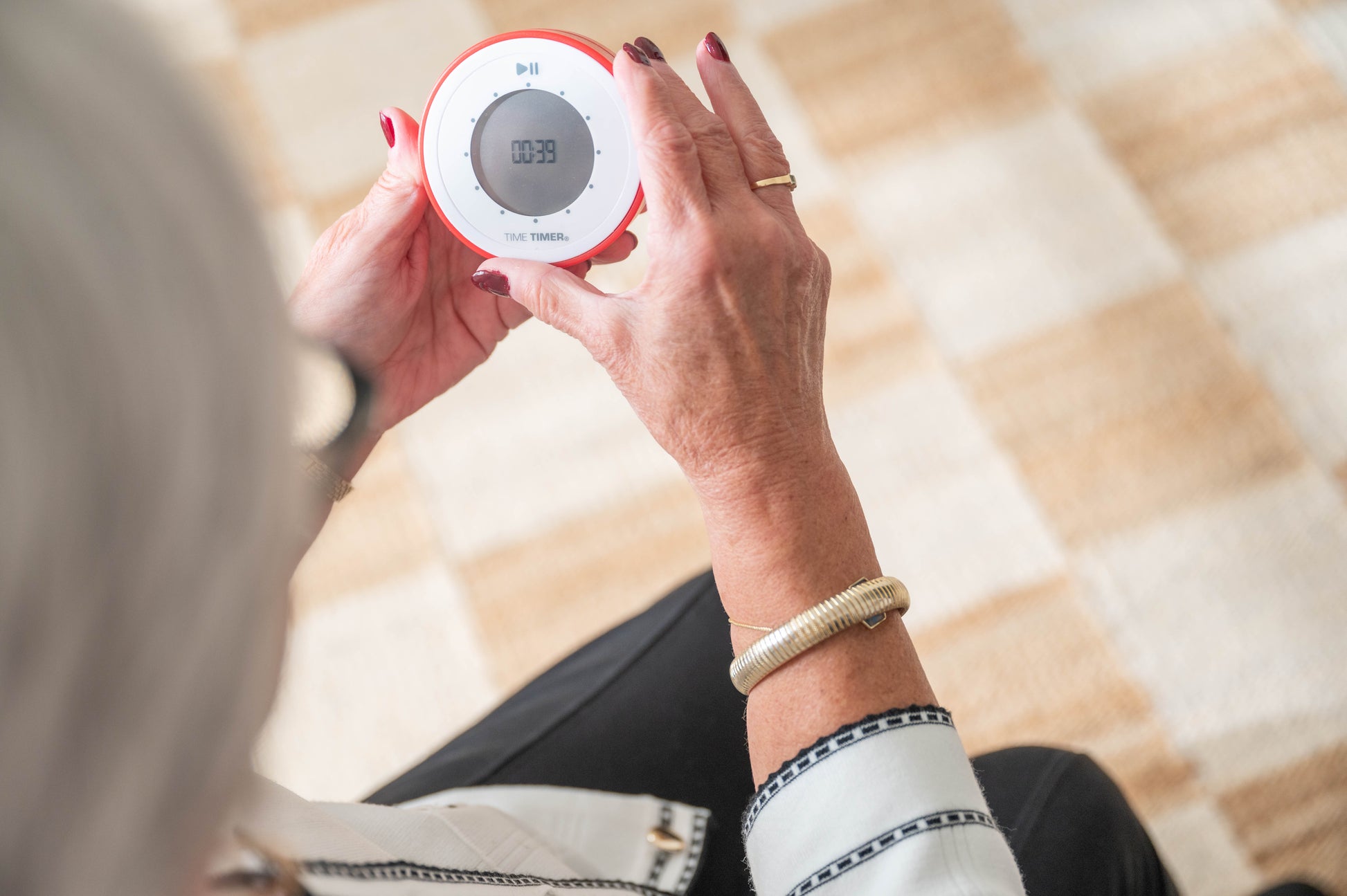 An older woman with painted nails holds a Time Timer Twist with Kickstand (Red) by Time Timer, showing 00:39 while seated on a patterned light brown and beige carpet.