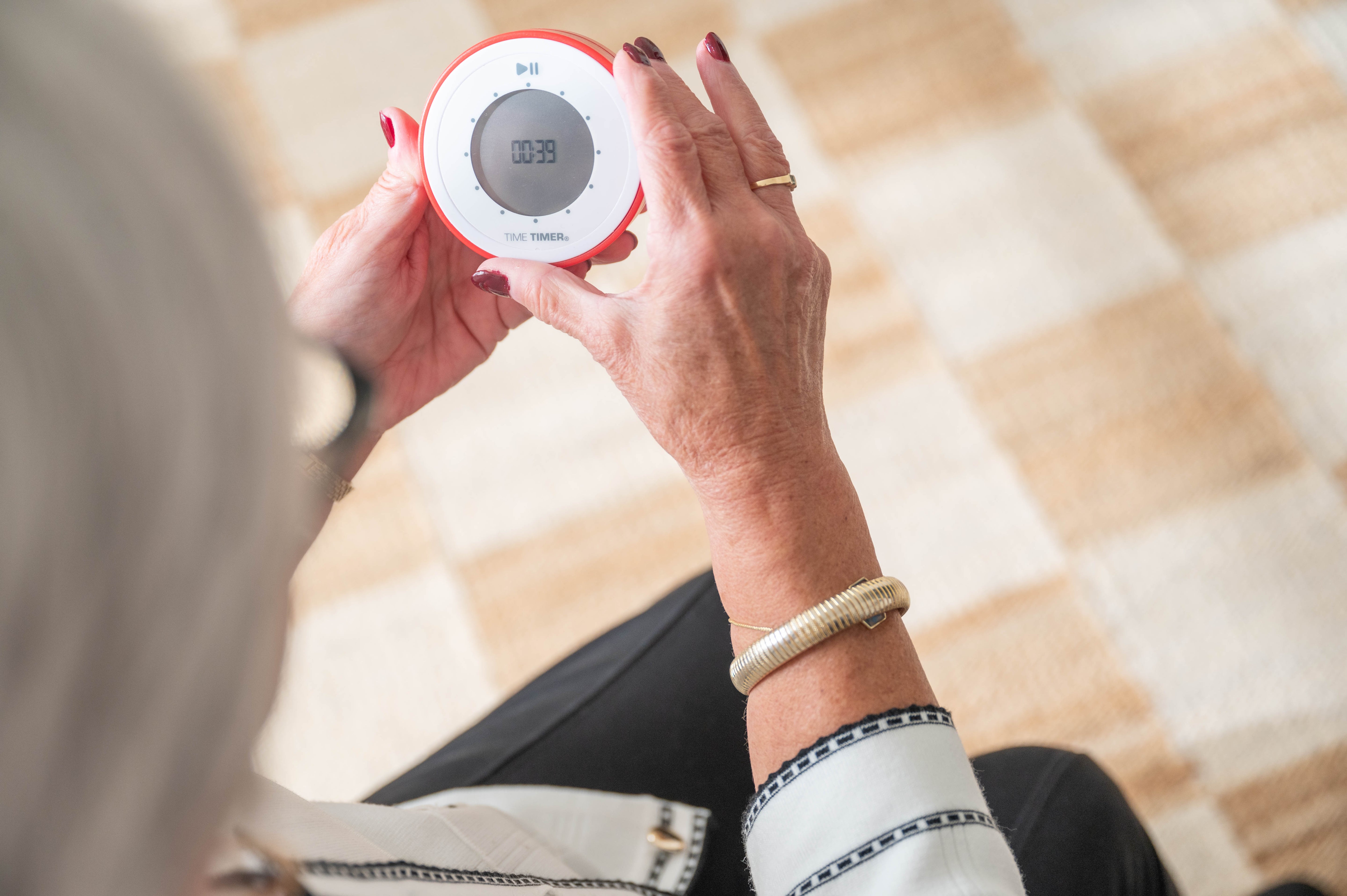 An older woman with painted nails holds a Time Timer Twist with Kickstand (Red) by Time Timer, showing 00:39 while seated on a patterned light brown and beige carpet.