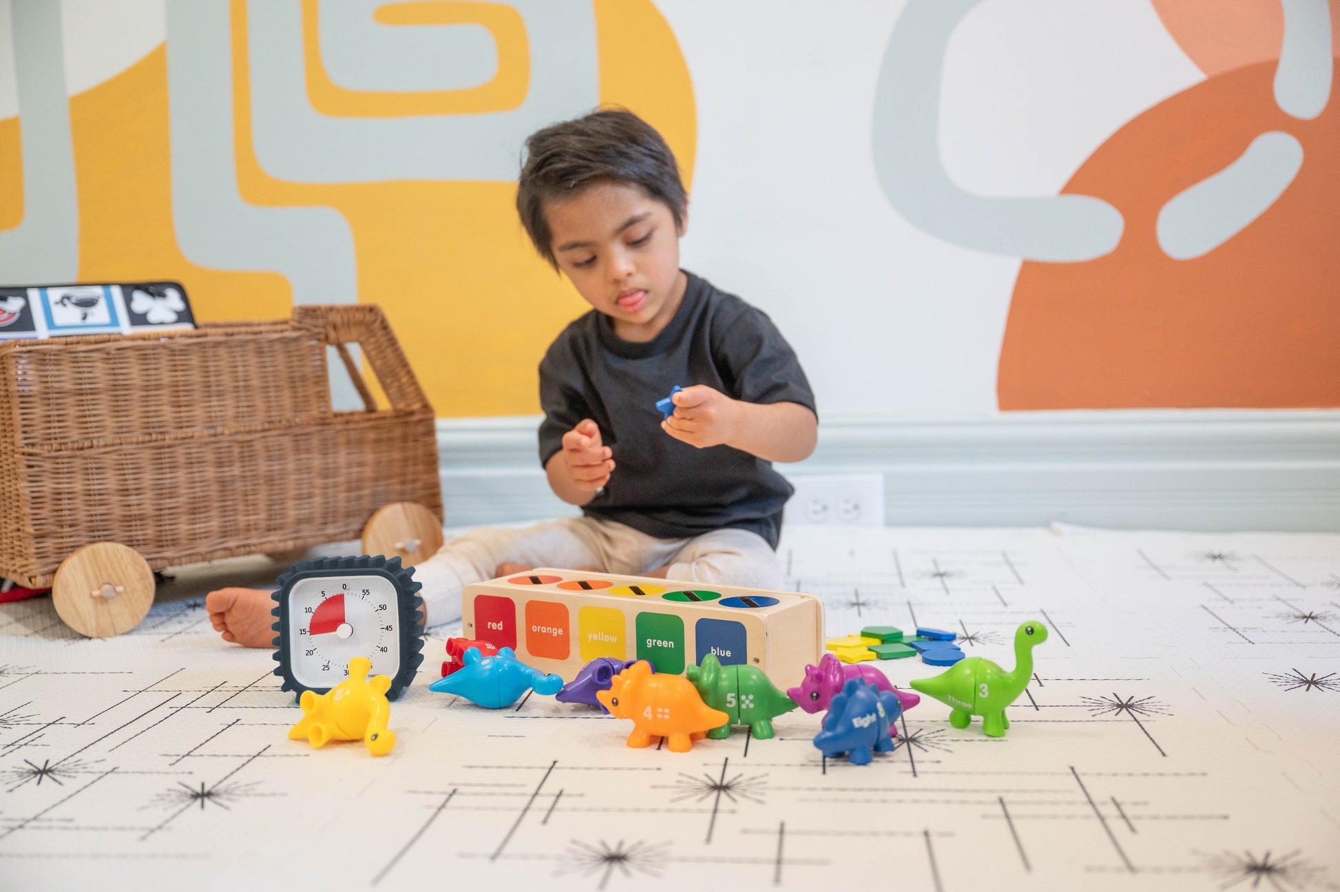 A young child plays with colorful dinosaur toys and a sorting block as the Time Timer MOD + Protect Case – Tread (60 min) by Time Timer tracks playtime nearby, set against a wall with bold, abstract designs.