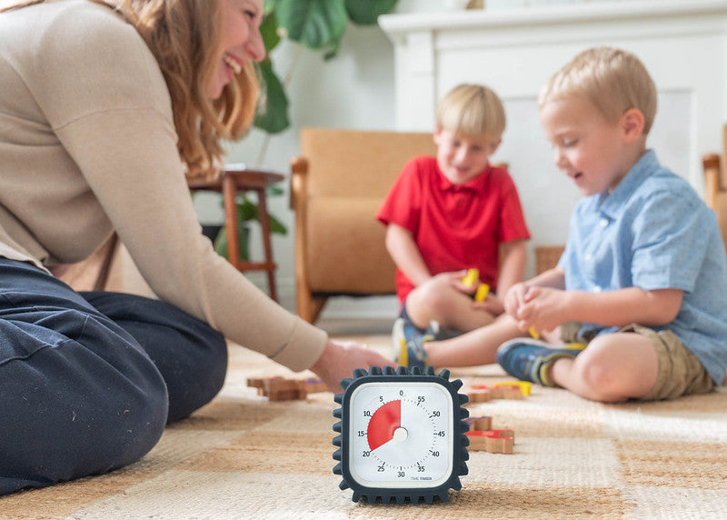 A Time Timer MOD + Protect Case – Tread (60 min) by Time Timer sits on a rug, while a smiling adult and two kids play with toys nearby in a bright, cheerful room that suggests a playful learning space.