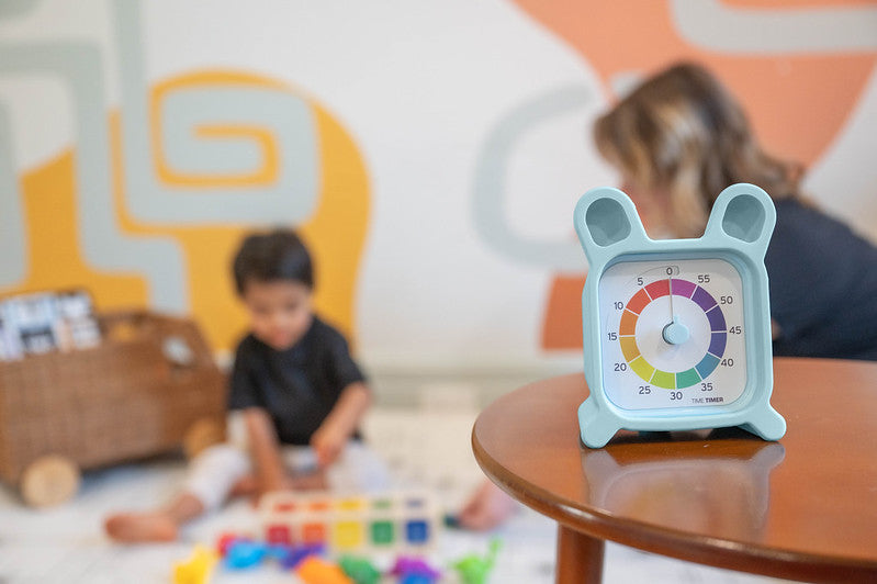 A Time Timer MOD + Protect Case – Bunny (60 min) by Time Timer, with its blue design and bunny silicone cover, rests on a table as a child plays nearby and an adult appears in the blurred background.