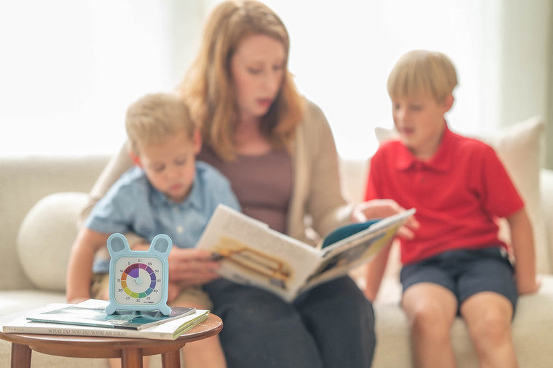 A woman reads to two boys on a sofa. On the table is a Time Timer MOD + Protect Case – Bunny (60 min) by Time Timer, featuring colored sections. The softly lit scene feels relaxed.