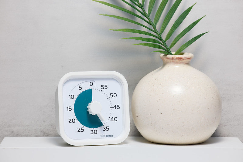The Time Timer PRO Applied Visual Timer (60 min) by Time Timer, shown in white with a blue dial set to 35 minutes, sits beside a cream ceramic vase with a palm leaf—perfect for medication reminders and memory care routines.