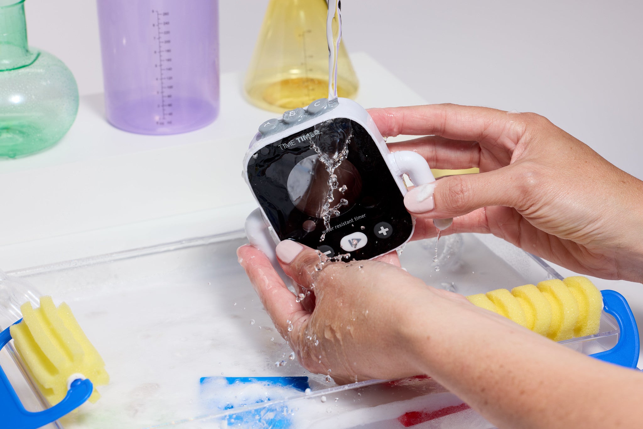 Someone washes a Time Timer Element by Time Timer under running water in a tray with soap, sponges, and cleaning tools. Colorful laboratory flasks are visible in the background.