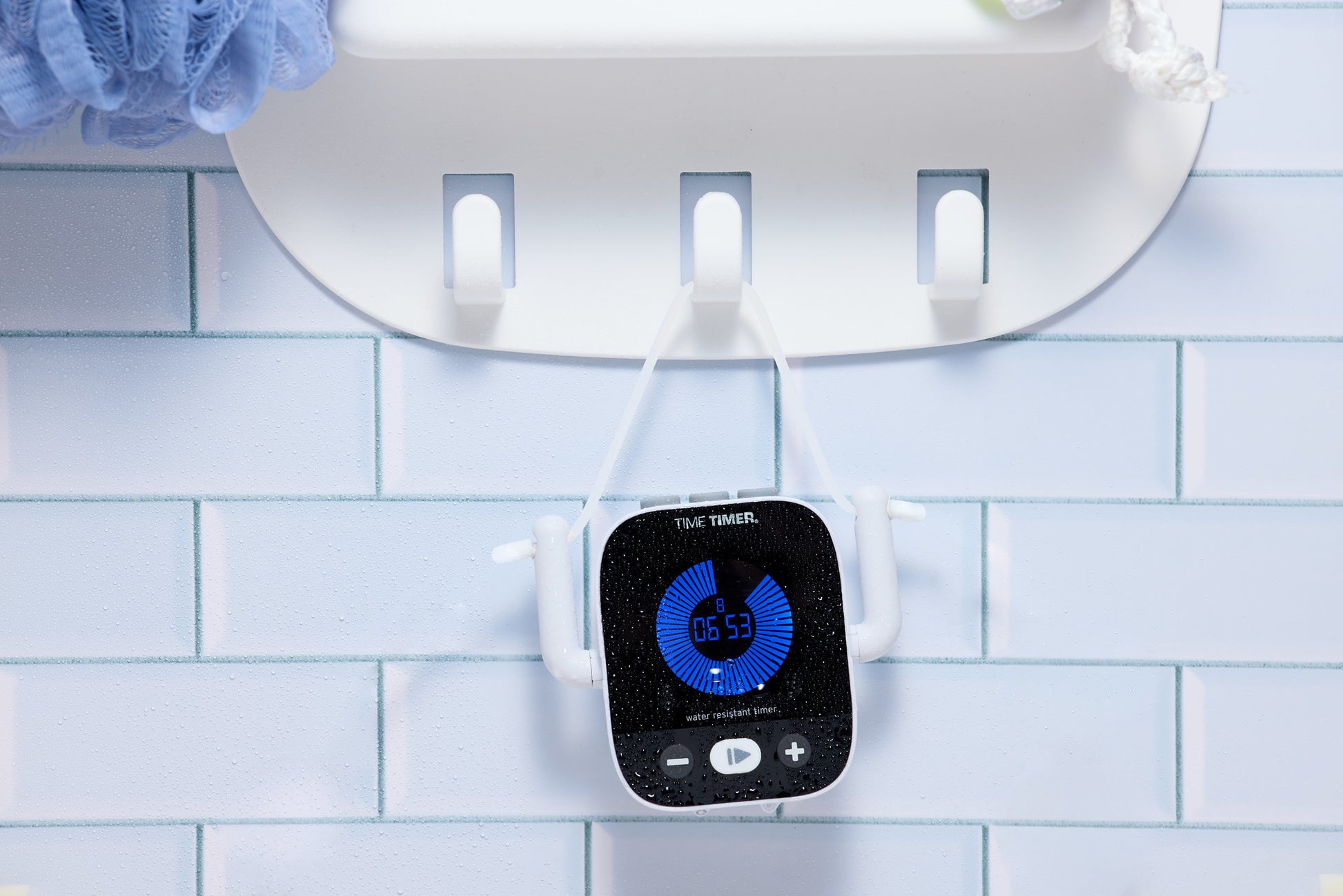 The Time Timer Element by Time Timer, a black water-resistant timer with a blue display, hangs from a white hook on a tiled bathroom wall. The timer reads 6:53 and has water droplets on its surface.