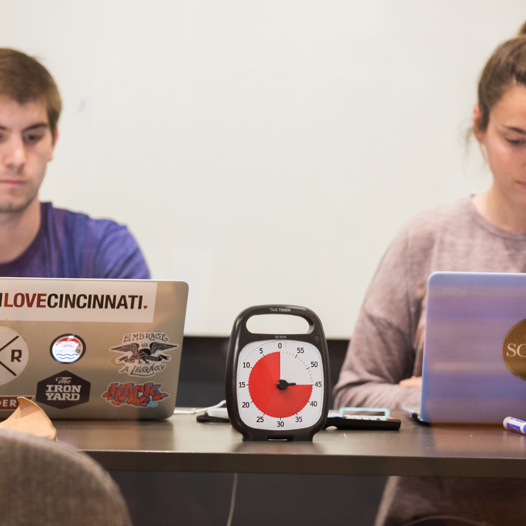 Two people work on laptops at a table, using the Time Timer Plus (60 min) by Time Timer to stay focused. Markers and erasers are scattered nearby, and a whiteboard is in the background for time management support.