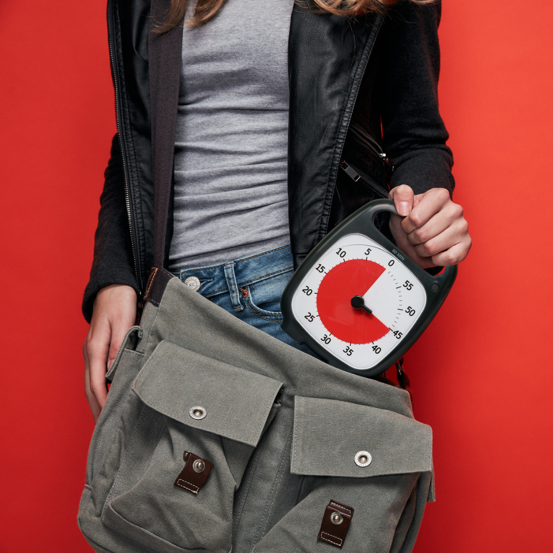 A person in a gray shirt and black jacket holds a Time Timer Plus (60 min) by Time Timer and a green canvas bag against a red background. The timer is partly inside the bag, showing its red countdown section.