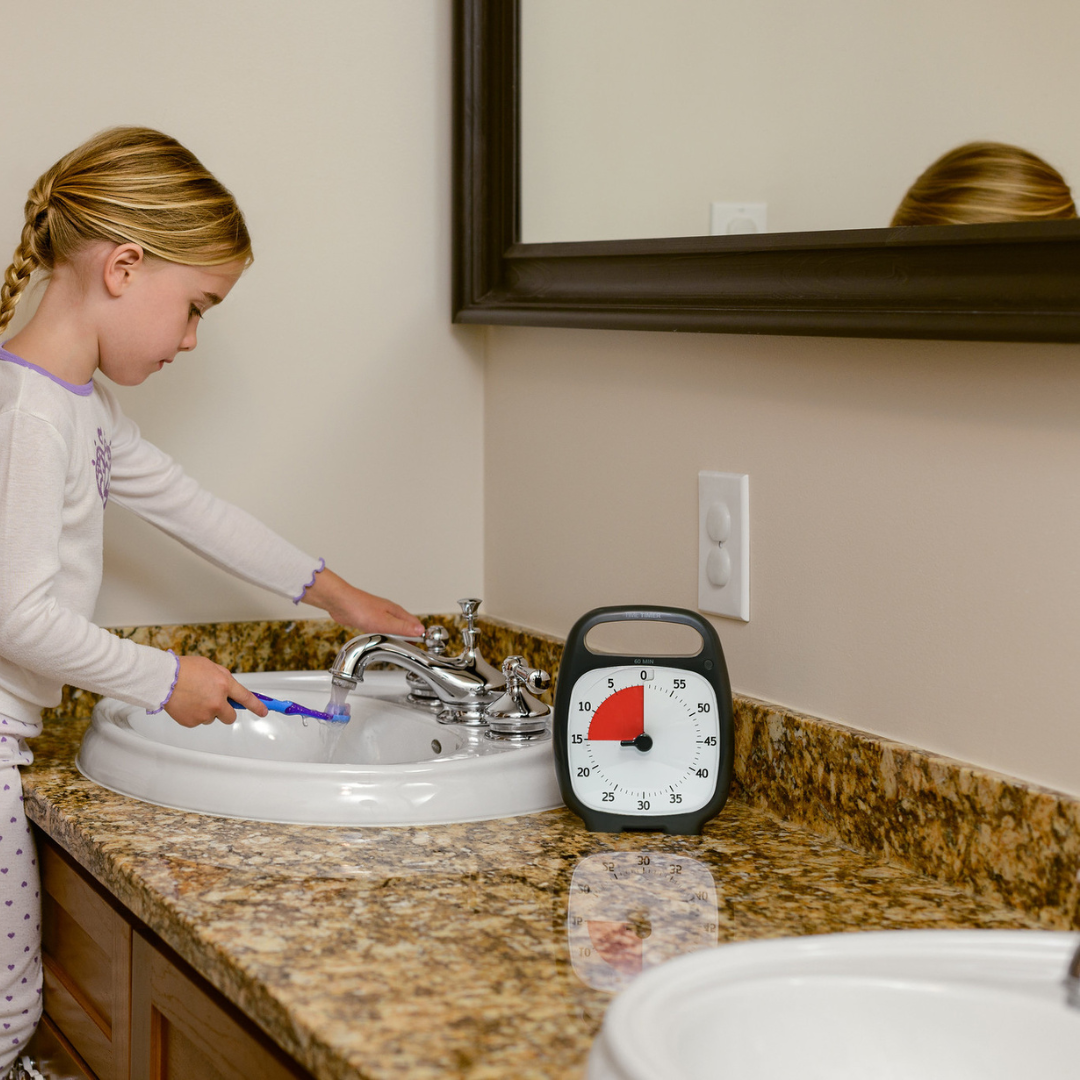 A young girl in pajamas brushes her teeth at the bathroom sink, with a Time Timer Plus (60 min) by Time Timer showing red time remaining on the counter beside her to help manage time.