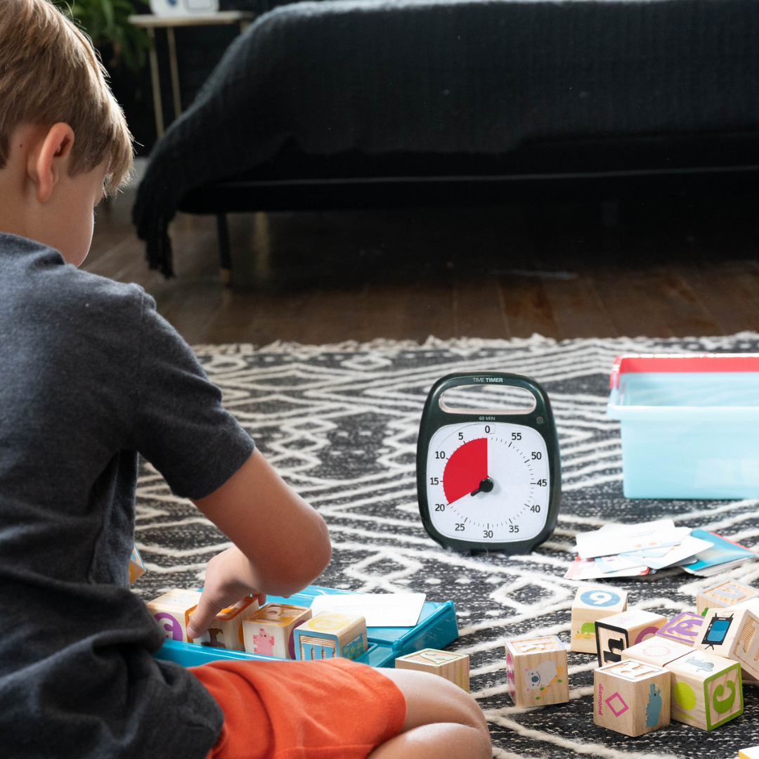 A child plays with colorful wooden blocks on a patterned rug near the Time Timer Plus (60 min) by Time Timer, showing about 20 minutes left, alongside a blue storage bin in a cozy room.