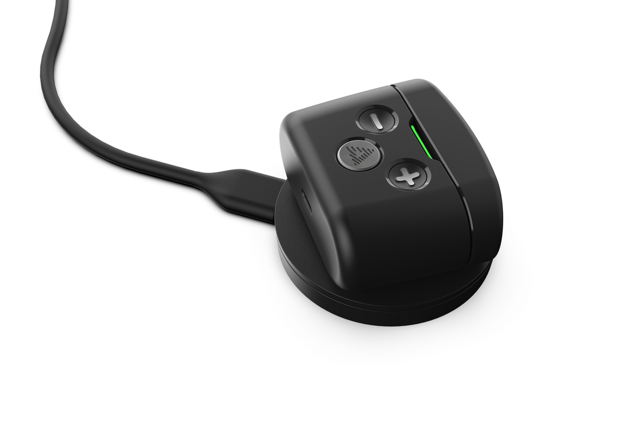 A close-up of black Audeara Buds Noise Cancelling Aid (In-Ear) with a mute button, volume control, and green LED indicator, connected to a charging cable and resting on its round charging base.