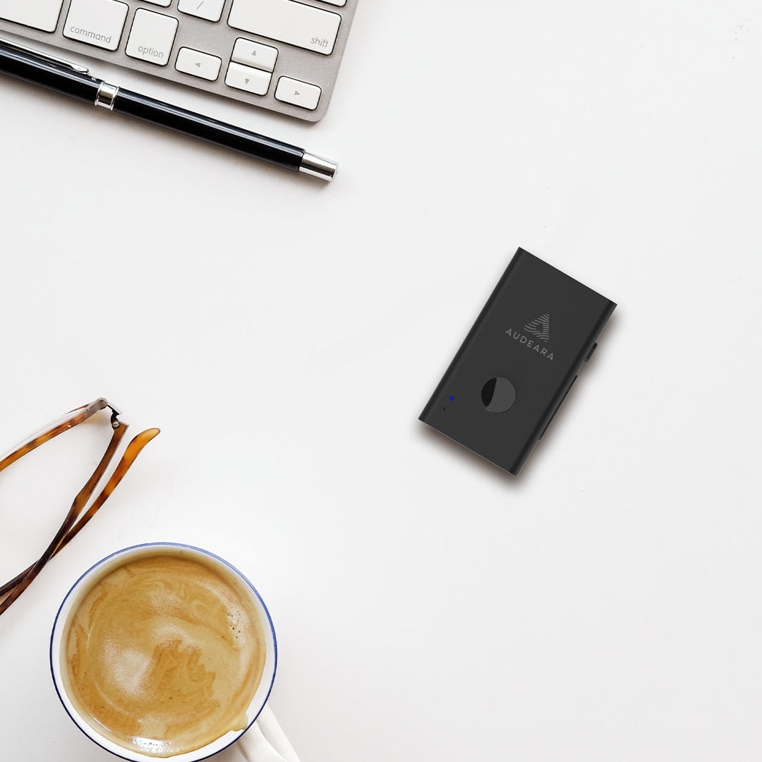 A cup of coffee, a pen, a keyboard, an Audeara Audio Transceiver & Lapel Microphone – Auracast™ & Bluetooth®, and a black A6 notebook labeled AQUAREA sit on a white desk.