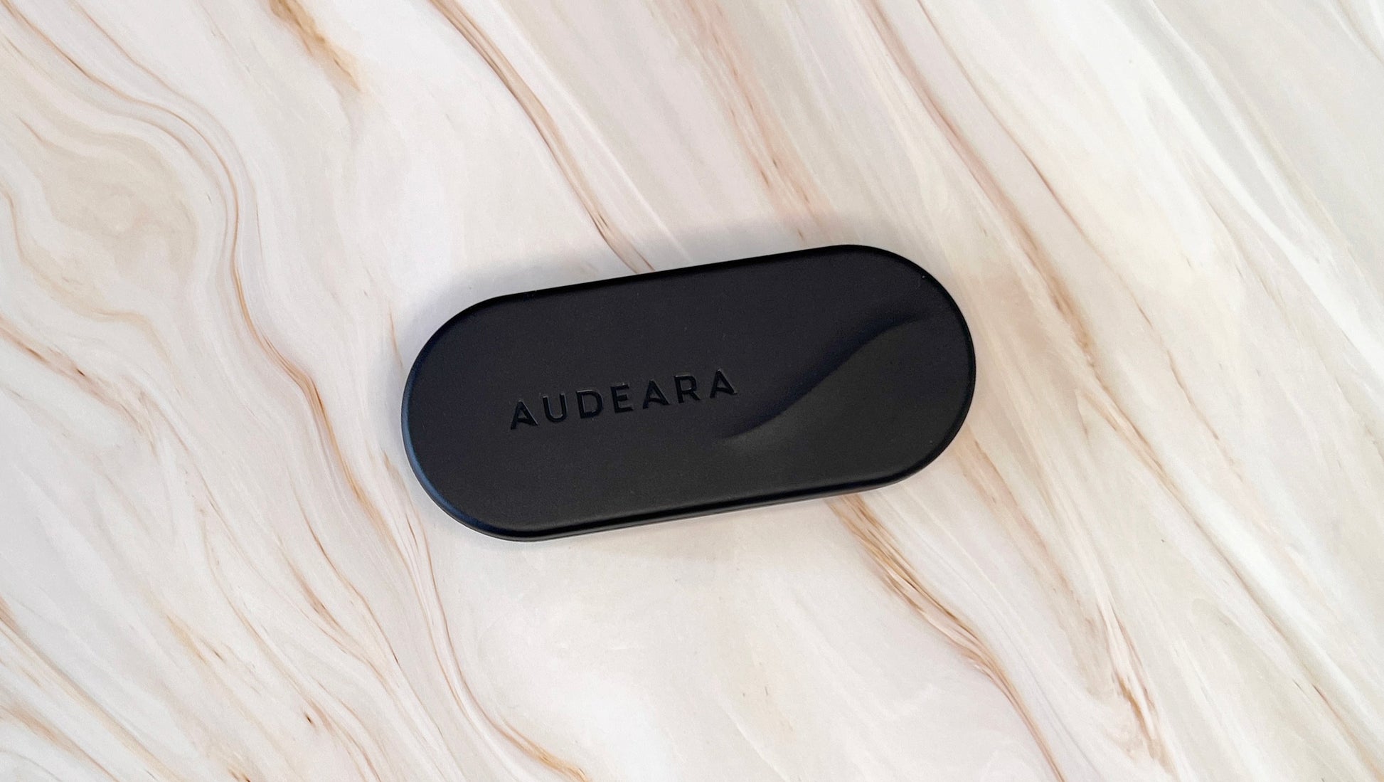 A black, rectangular Audeara Broadcaster (Auracast™ Transmitter) with rounded edges sits on a light marble surface veined in beige and brown, highlighting Audeara’s dedication to inclusive listening.
