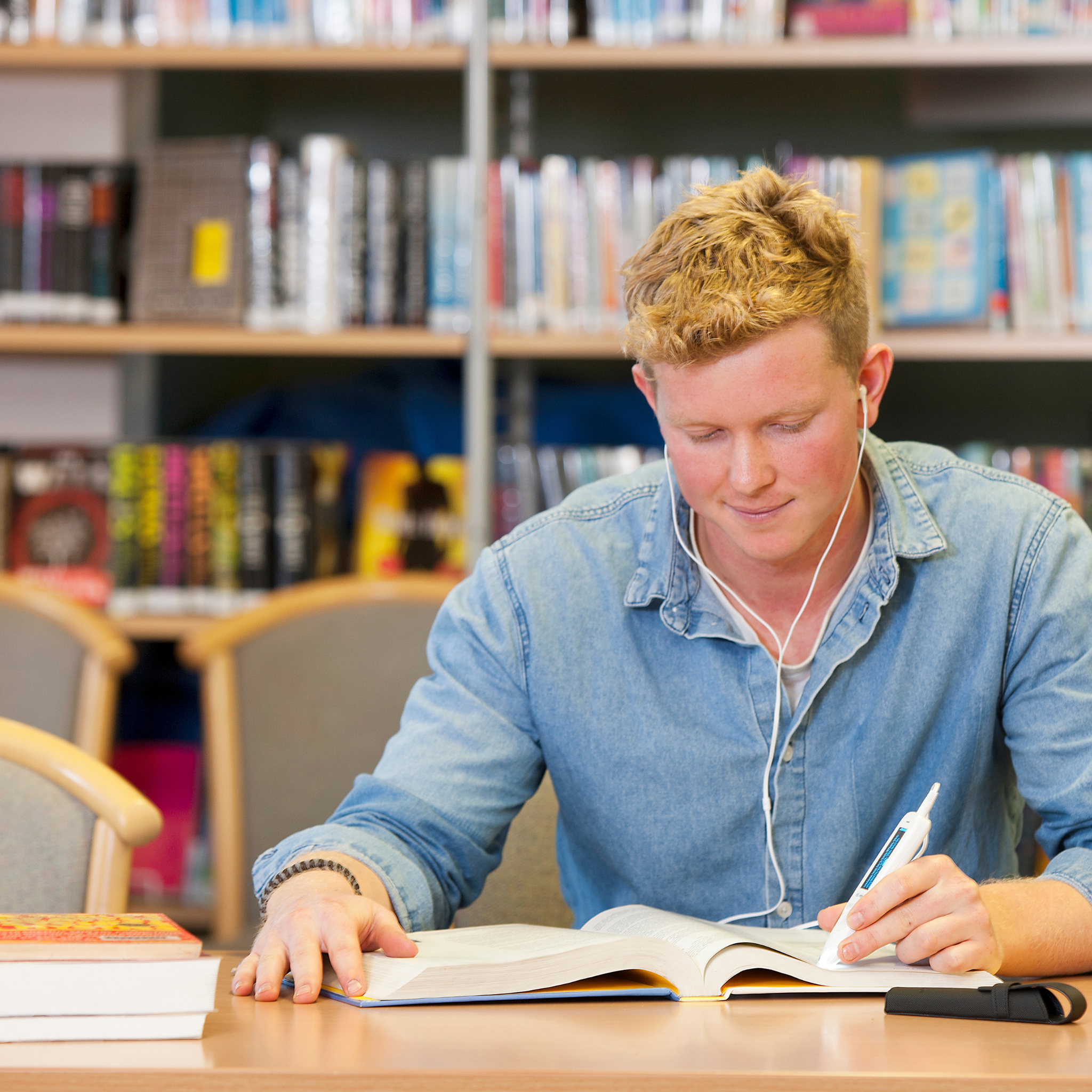 A young man in a light denim shirt studies at a library table, using the C-PEN C-Pen Reader 2 to write in an open book. He wears earbuds, and bookshelves filled with colorful books are visible in the background.