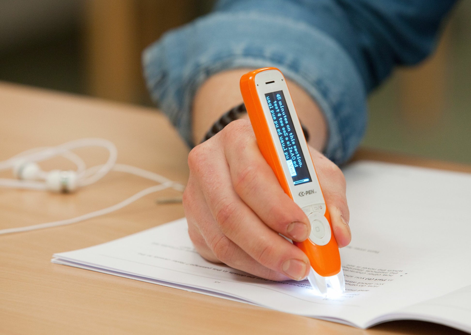 A person uses a C-PEN C-Pen Exam Reader 2 in orange to scan text from a printed document; the device's screen shows the text, while earphones and a blue sleeve are visible in the background, showcasing this assistive reader.