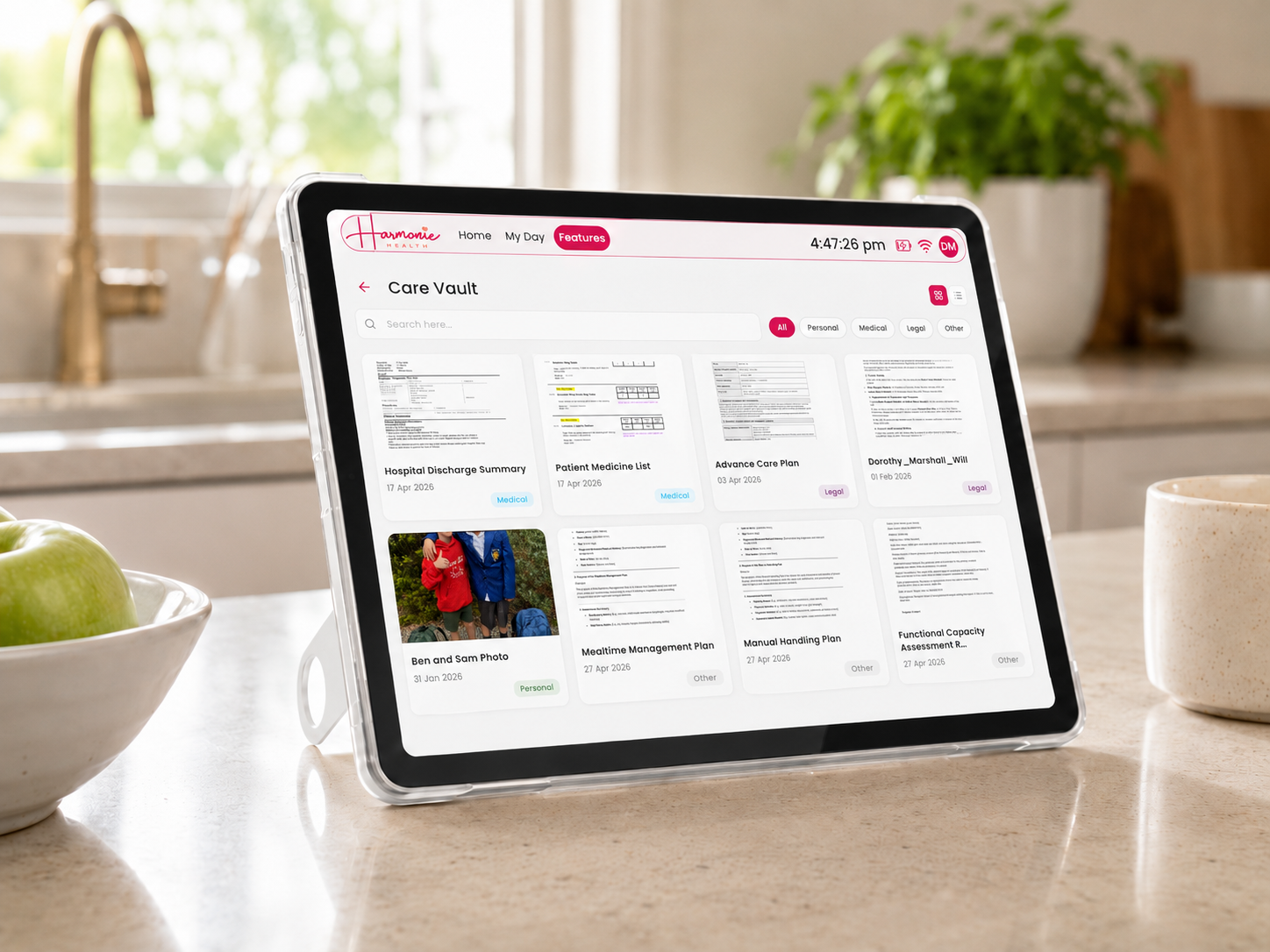 A Harmonie Health Hub with Subscription by Harmonie Health sits on a kitchen counter, showing organized medical and personal files on its screen. A bowl of green apples and a beige mug are nearby.