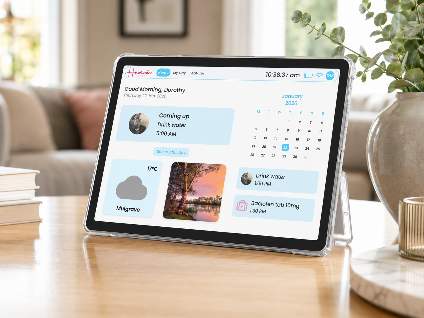The Harmonie Health Hub by Harmonie Health displays your daily schedule, medication and hydration reminders, weather, photos, calendar, and time. It sits on a wooden desk beside books, a mug, and a plant. Subscription required.