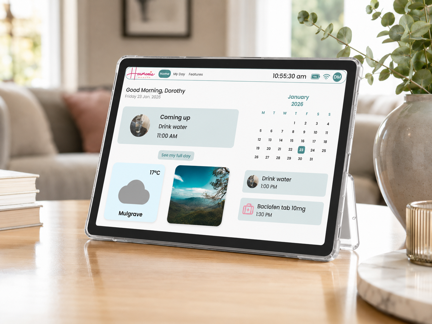 The Harmonie Health Hub with Subscription by Harmonie Health sits on a wooden table, displaying a digital dashboard showing date, time, weather, events, reminders, and calendar. Books, a sofa, and a plant appear in the background.