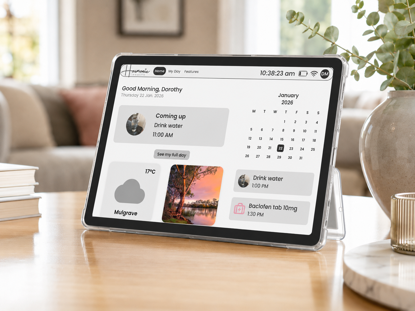 The Harmonie Health Hub with Subscription by Harmonie Health is shown on a stand, displaying a personal dashboard with date, time, weather, calendar, to-dos, medication reminders, and a scenic photo. It's placed on a wooden table beside books and a plant.