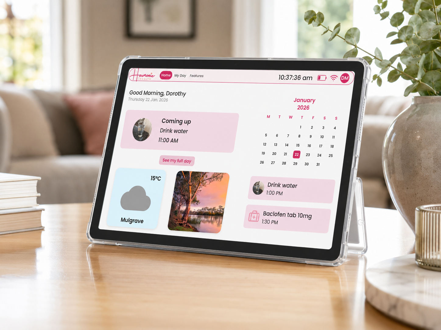 The Harmonie Health Hub with Subscription by Harmonie Health sits on a table displaying reminders, a weather update, the January 2026 calendar, and photos. In the background are a plant, lamp, and books.