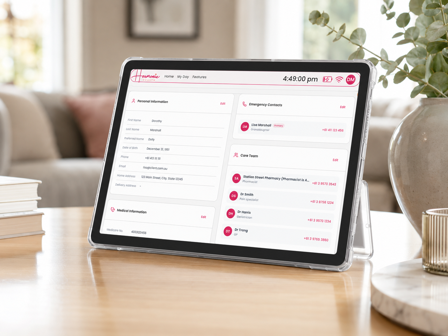 A Harmonie Health Hub with Subscription tablet by Harmonie Health sits on a table, displaying a health app interface with personal info, emergency contacts, care team, and medical details. Books, a plant, and a blurred background complete the scene.
