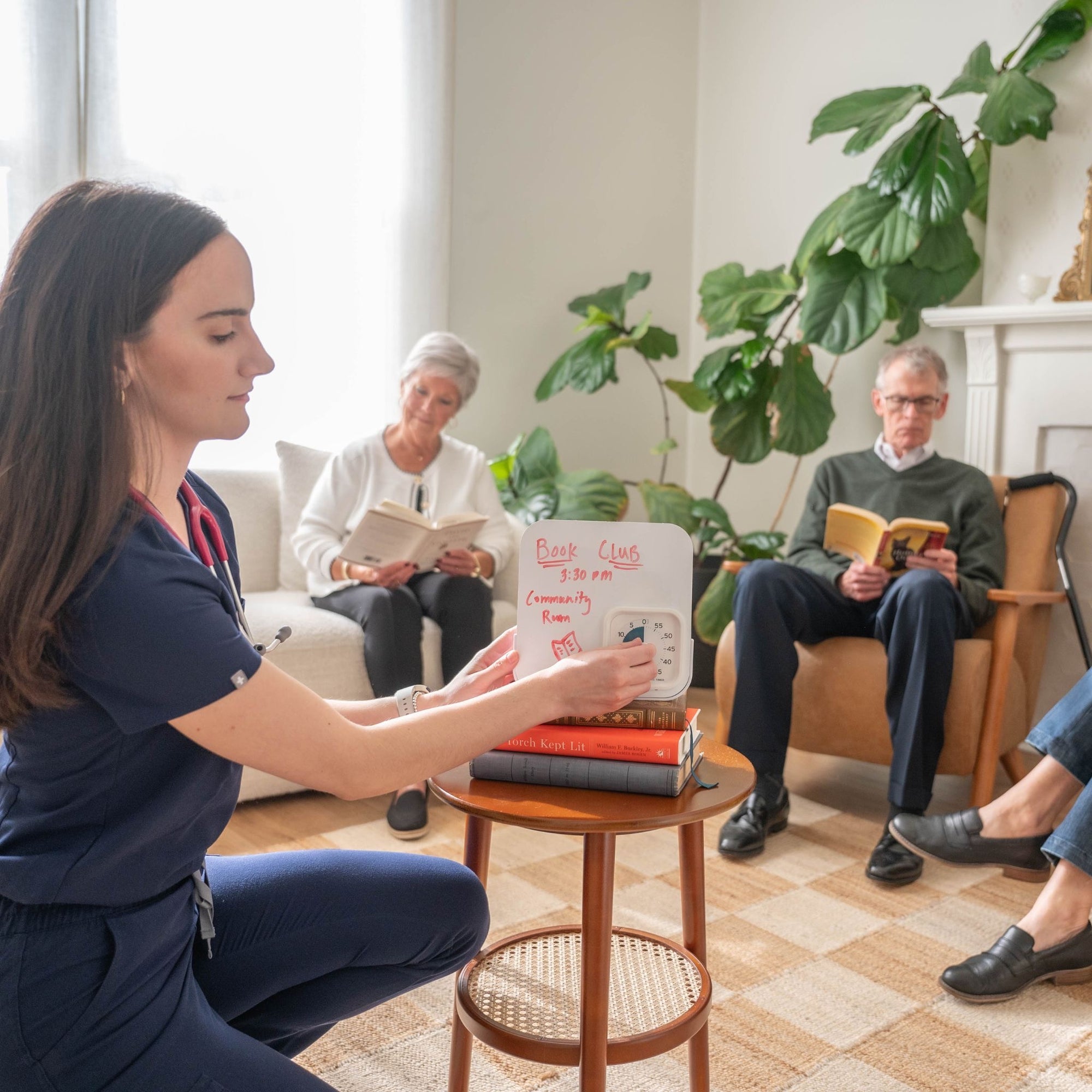 A woman in scrubs writes on a small whiteboard labeled Book Club while three seniors sit in a bright living room, reading books. A large plant and a fireplace are in the background.