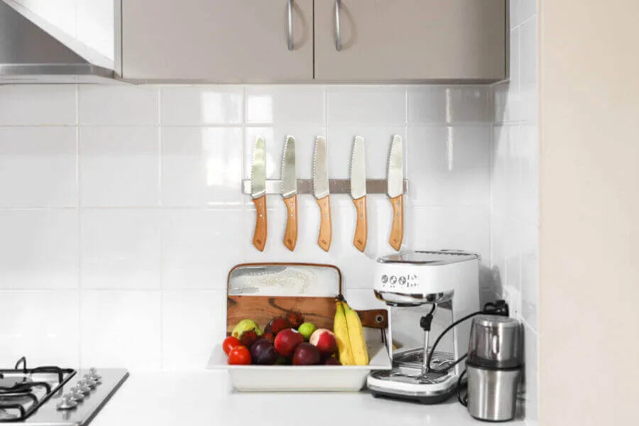 A modern kitchen countertop displays a bowl of fruits, bread box, coffee machine, grinder, and five KiddiKutter KandoKutter Adult Safe Knives [Wooden] mounted on a magnetic strip against a white tiled backsplash.