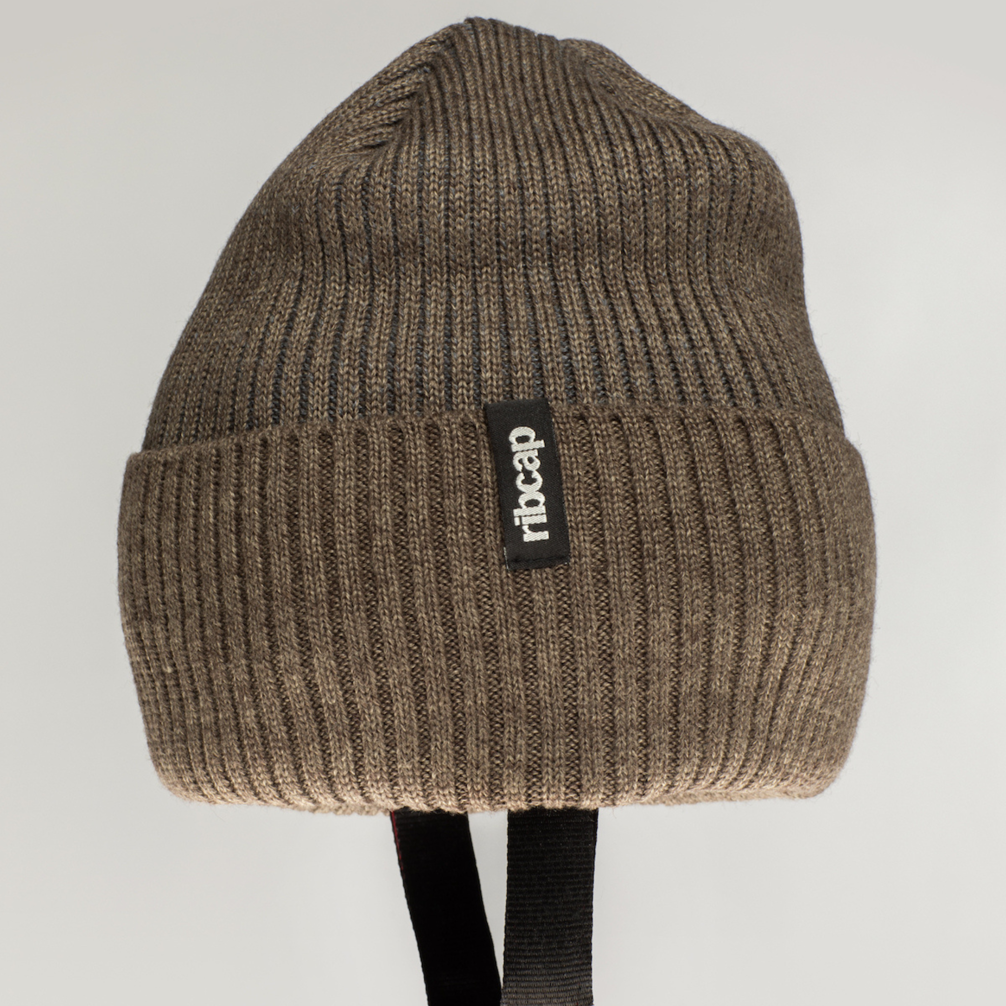 The Ribcap Iggy Beanie Protective Helmet in brown features a ribbed knit with a folded brim, black Ribcap label, and two black chin straps. This soft helmet provides discreet comfort and safety. The background is plain and light-colored.