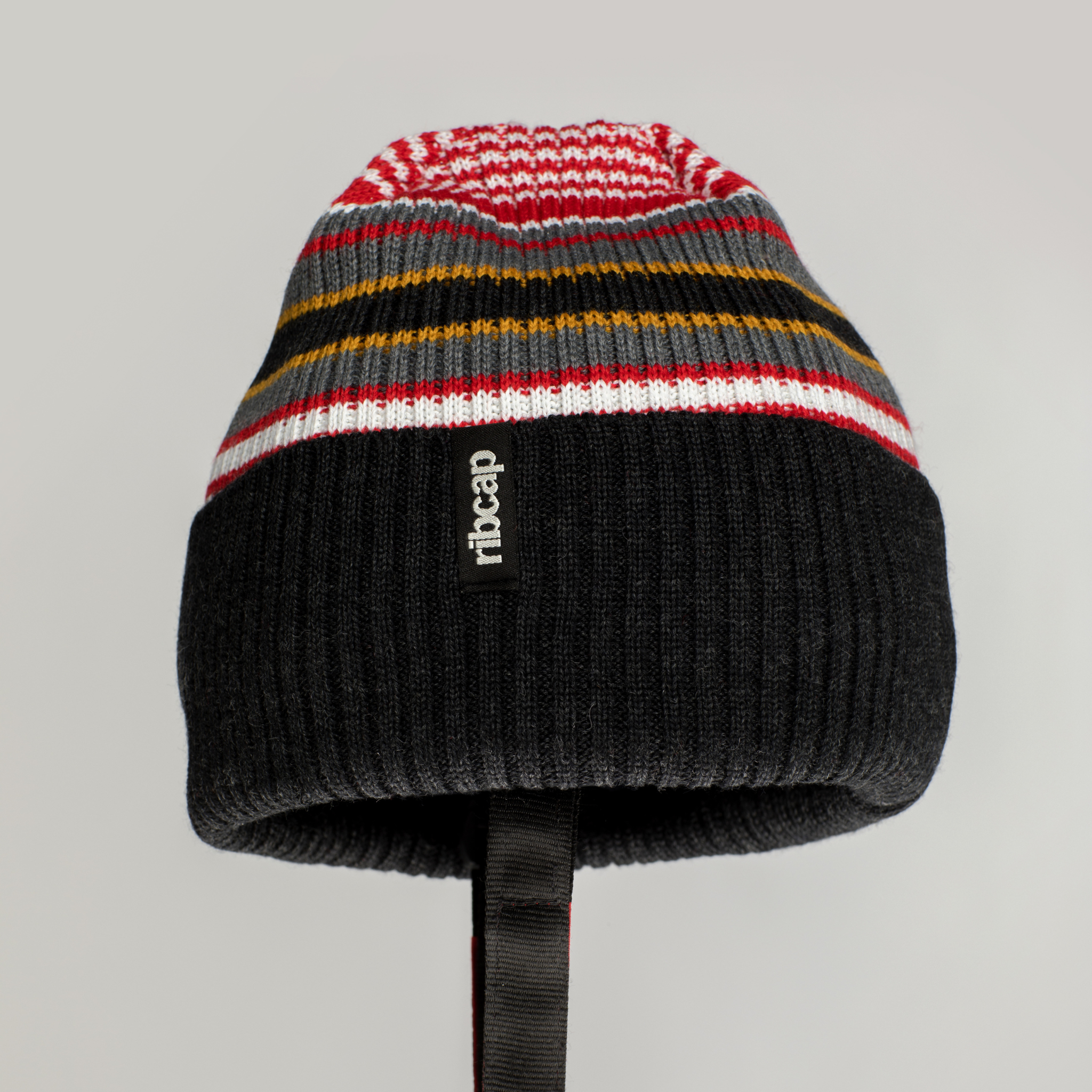 The Ribcap Iggy Beanie Protective Helmet [Stripy] by Ribcap is a soft, knitted helmet with black, gray, white, red, and yellow stripes, a folded brim with "ribcap" label, and a black strap for secure fit.