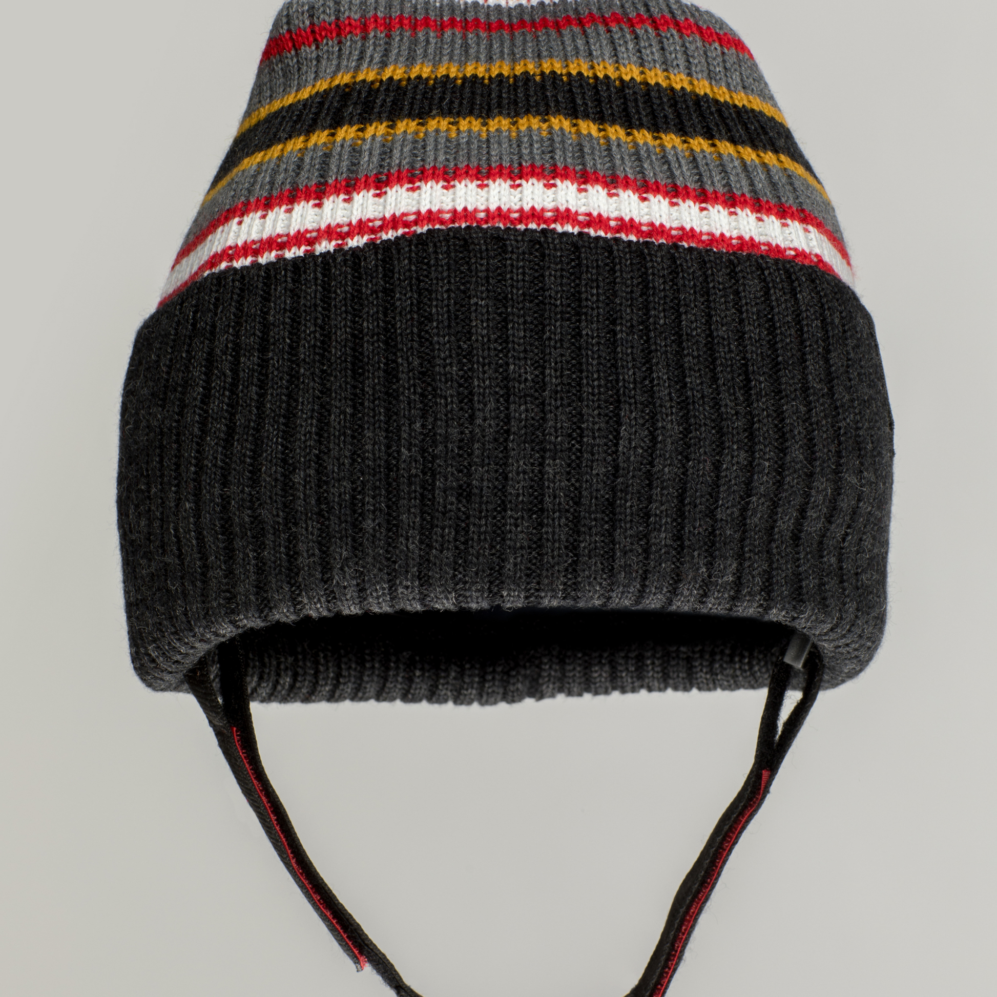 The Ribcap Iggy Beanie Protective Helmet [Stripy] by Ribcap is a helmet-style knitted winter hat with dark gray ribbing, multicolored stripes (yellow, red, white, gray), ear flaps, and chin straps for added warmth and comfort.