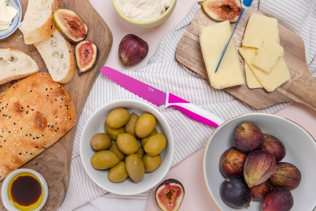 A spread of sliced bread, green olives in a bowl, cut figs, cheese slices on a wooden board, a purple knife, and a bowl of whole figs on a light table with a striped napkin.